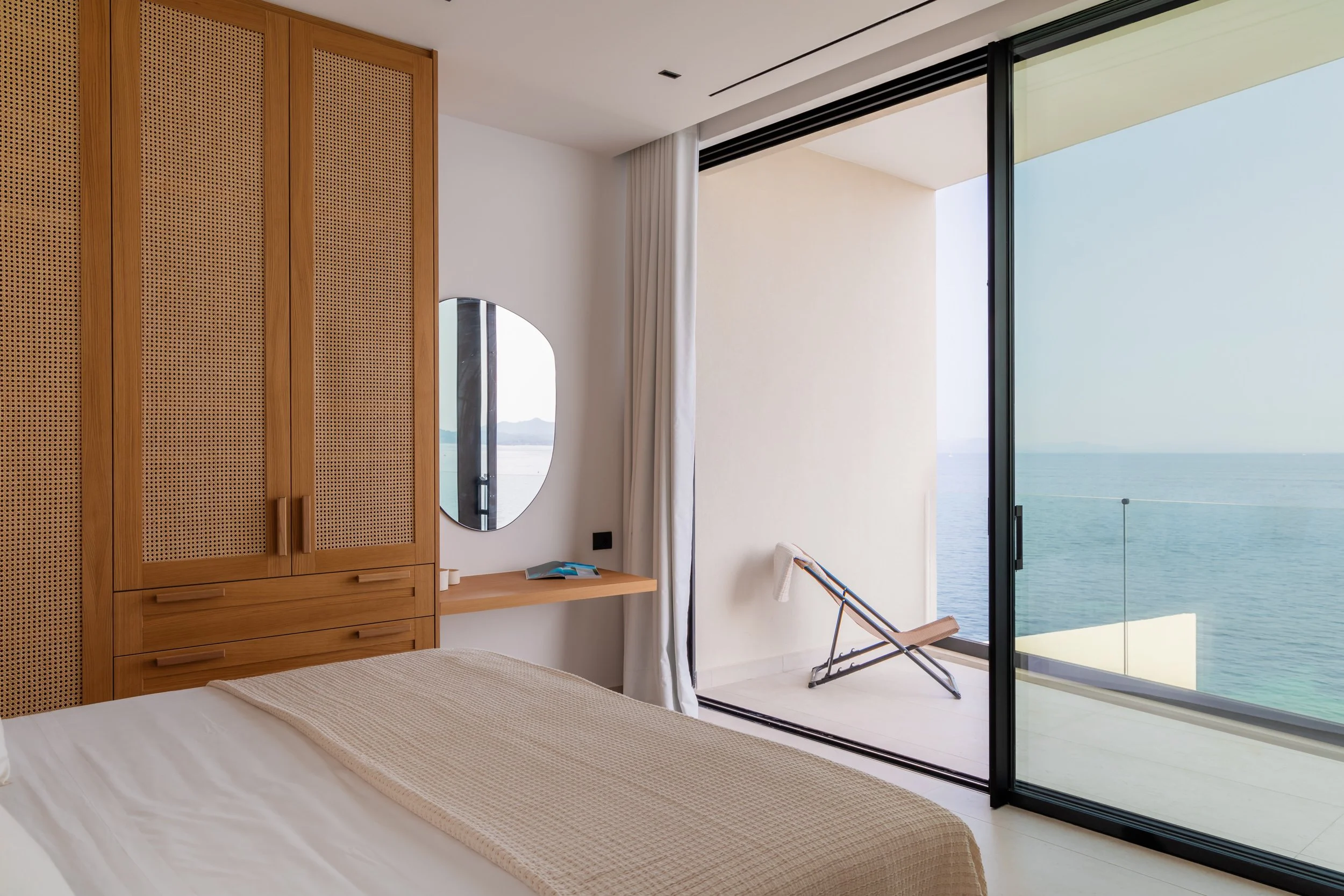 Modern bedroom with a large glass sliding door opening to a balcony overlooking the ocean, a wooden wardrobe, a mirror, and a bed with a beige blanket.