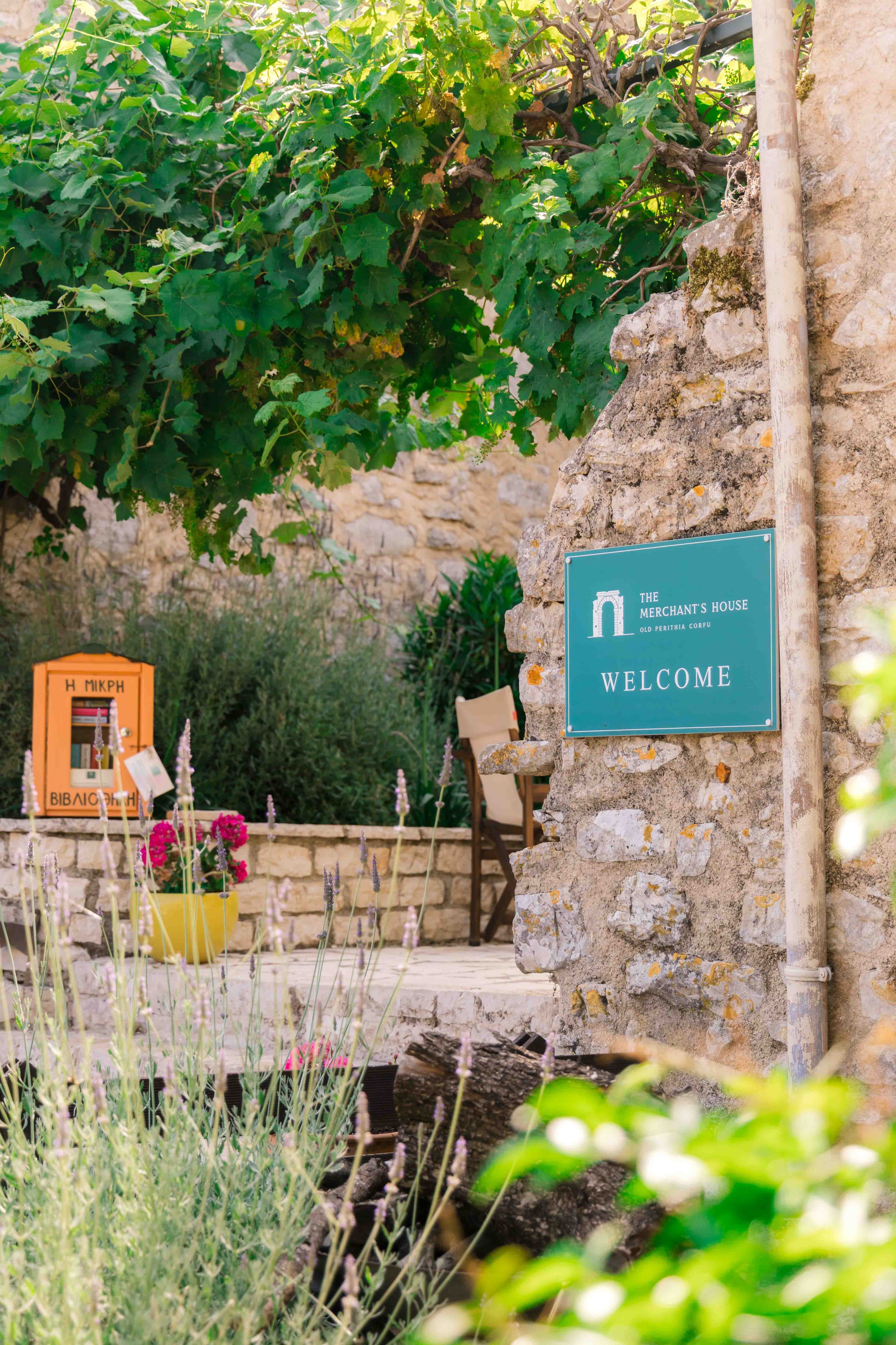 A garden scene with a stone wall, green vine-covered arbor, purple and pink flowers, a yellow pot, and a blue welcome sign on the wall that says 'The Merchant's House, Old Perithia Corfu, Welcome'.