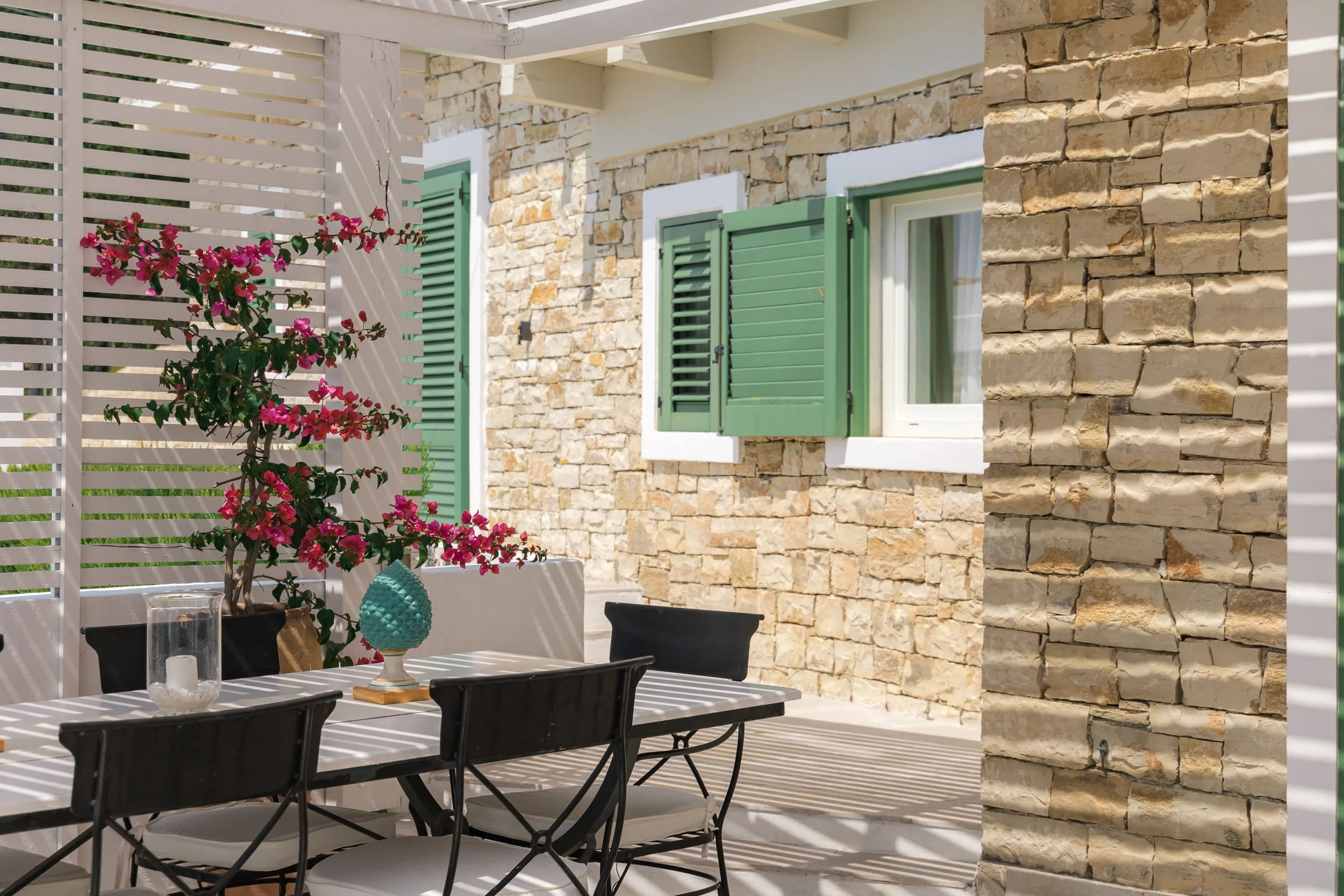 Outdoor dining area with a table and chairs, decorated with a turquoise vase and a clear glass candle holder, surrounded by a stone wall, green shuttered windows, and a pink flowering plant.