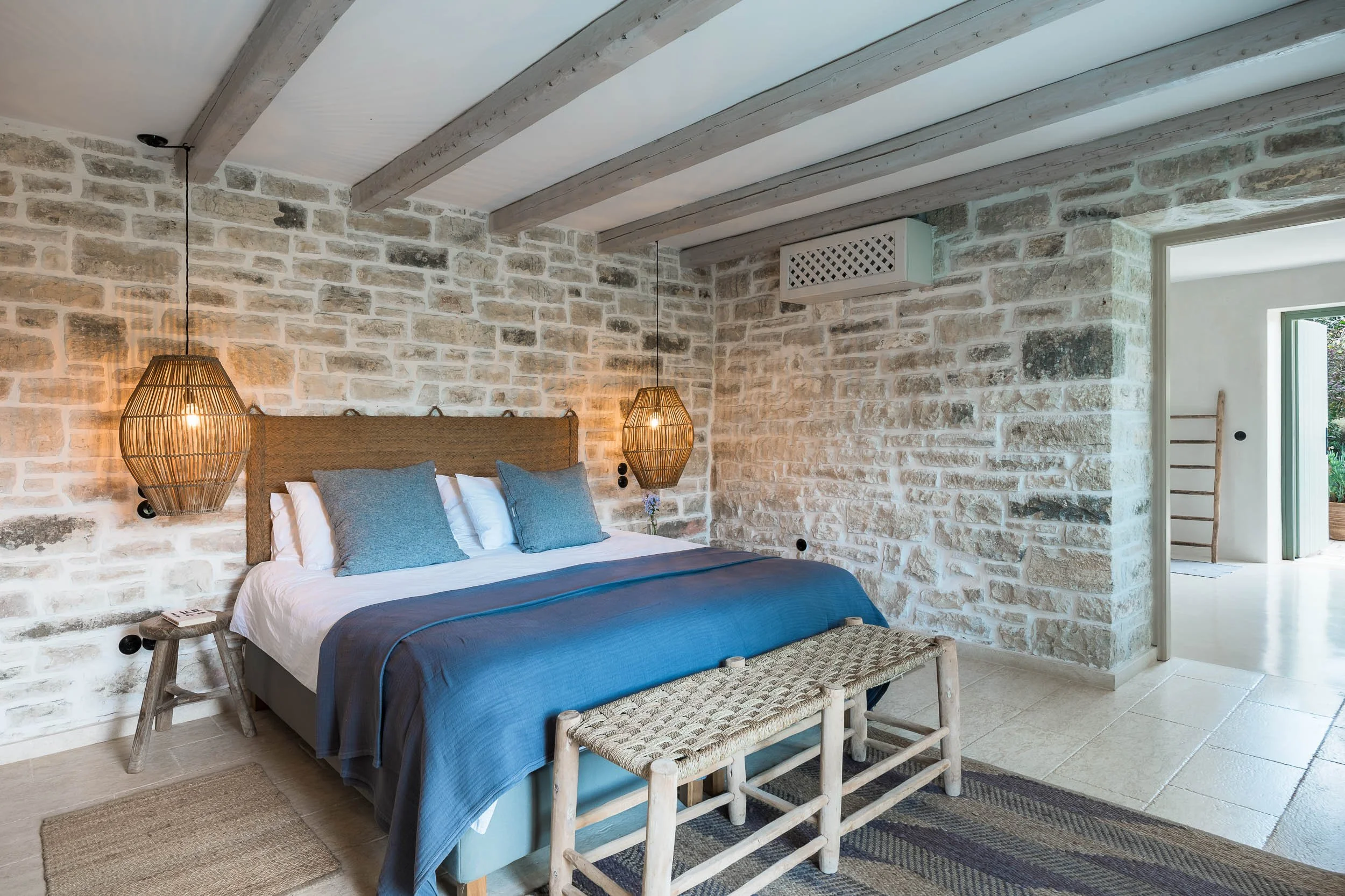 Bedroom with stone brick wall, wooden beams on ceiling, twin pendant lights, bed with gray and white bedding, and a small woven bench at the foot of the bed.