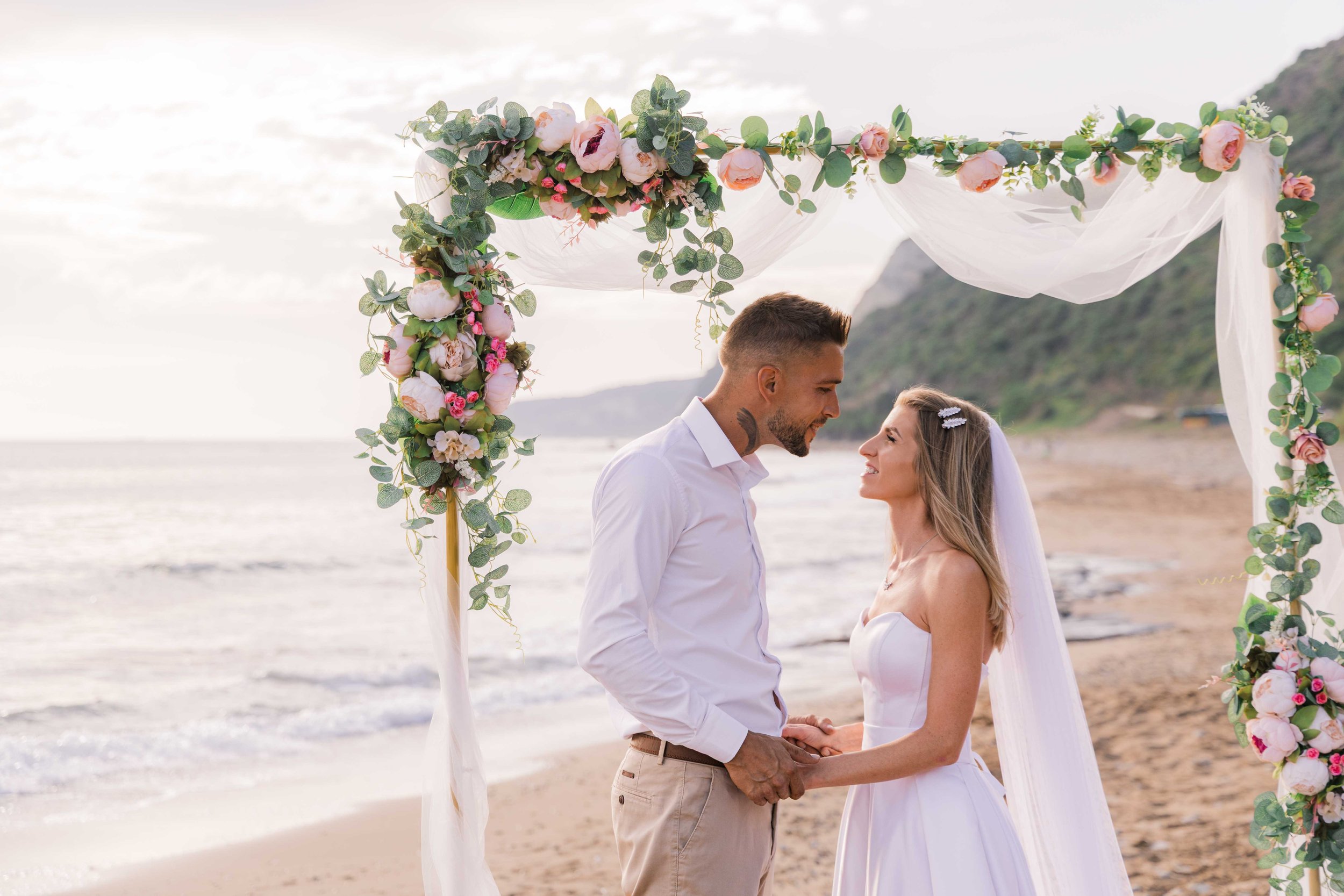 A couple getting married under an arch decorated with pink and white flowers and green foliage on a beach.