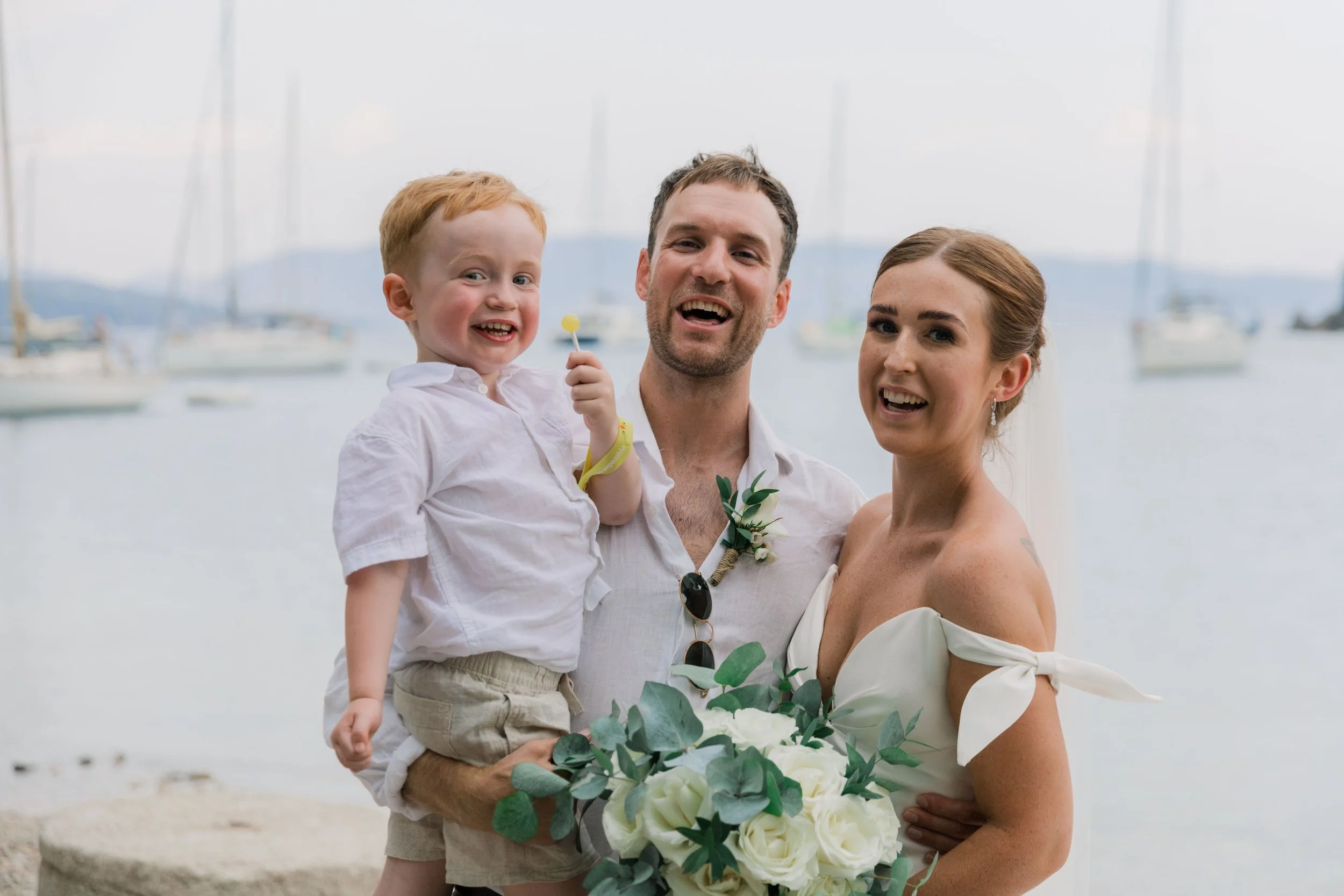 A happy family at a boat dock, with a man holding a young boy and a woman holding a bouquet of white roses, all smiling.