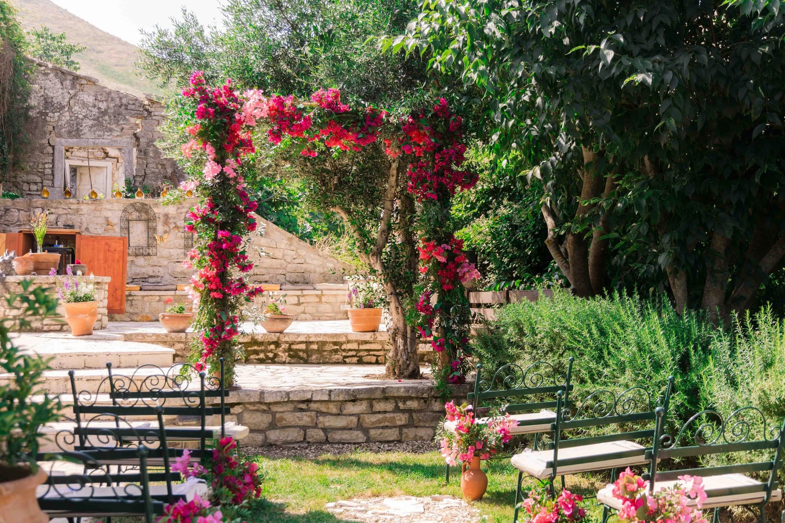 A garden with a stone patio, colorful flowering bougainvillea arch, potted plants, lush green trees, and iron benches with flower decorations.
