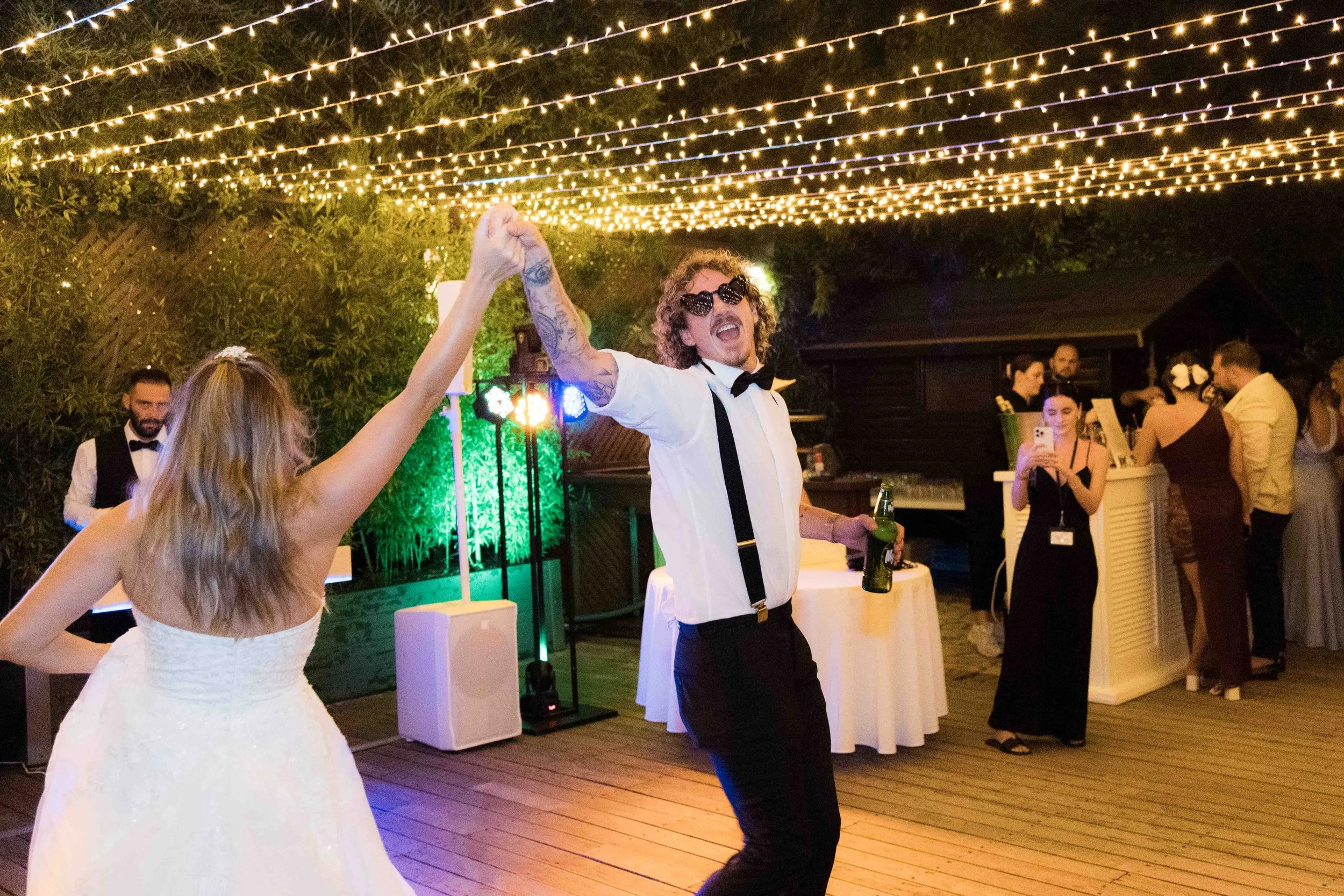 A joyful man and woman dancing at a wedding reception under string lights, with other guests in the background.