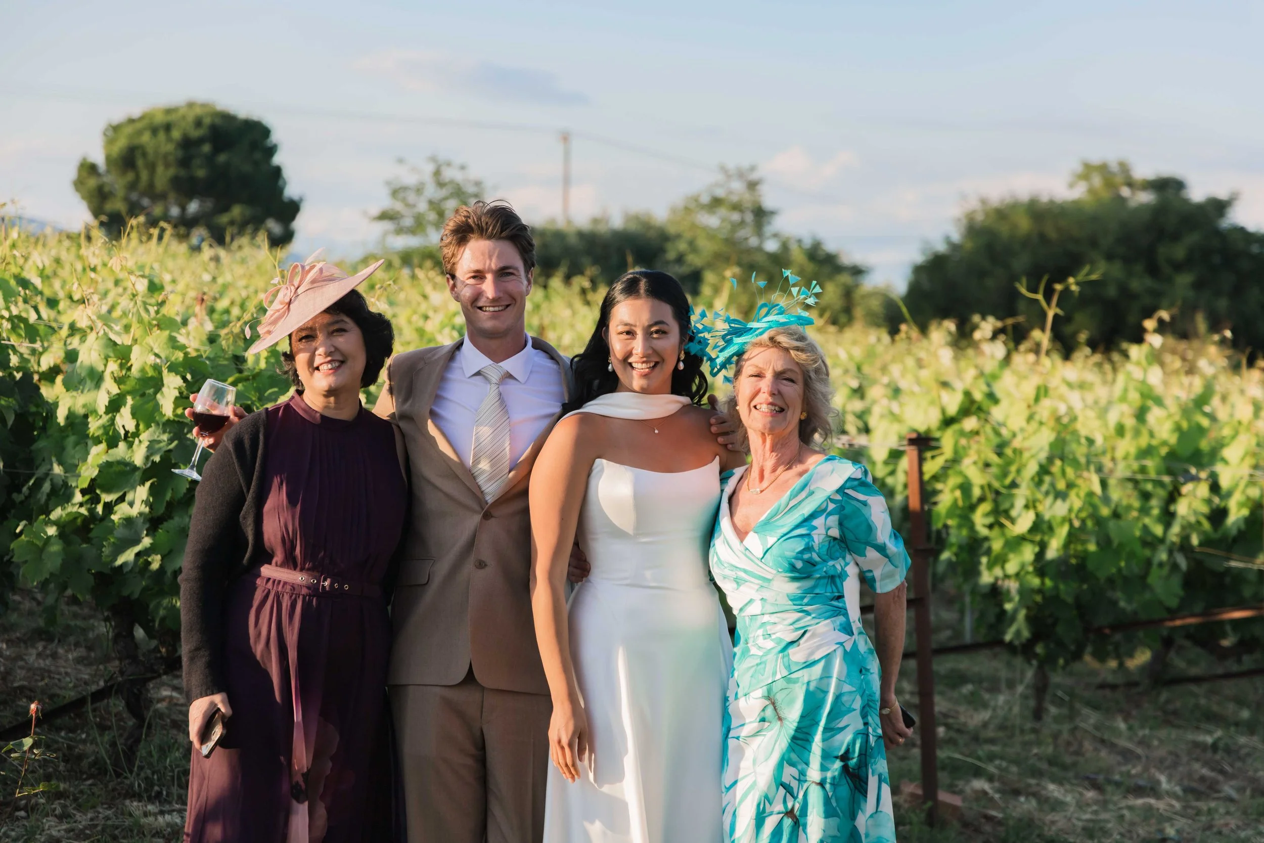 Group of five people dressed in wedding attire standing outdoors in a vineyard, smiling for a photo.