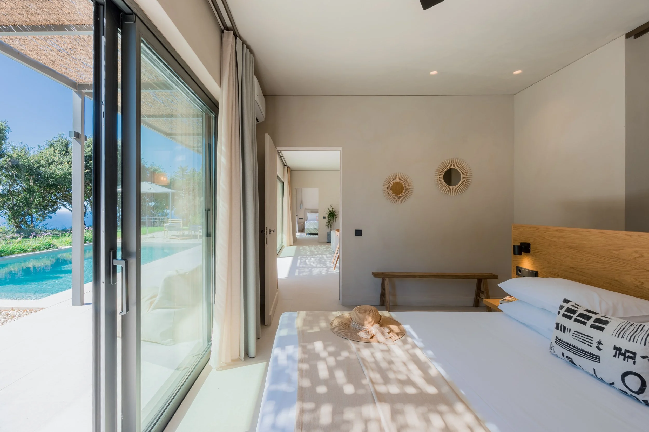 Sunlit hotel room with a bed, straw hat, and a pillow, open sliding glass door reveals a pool outside.