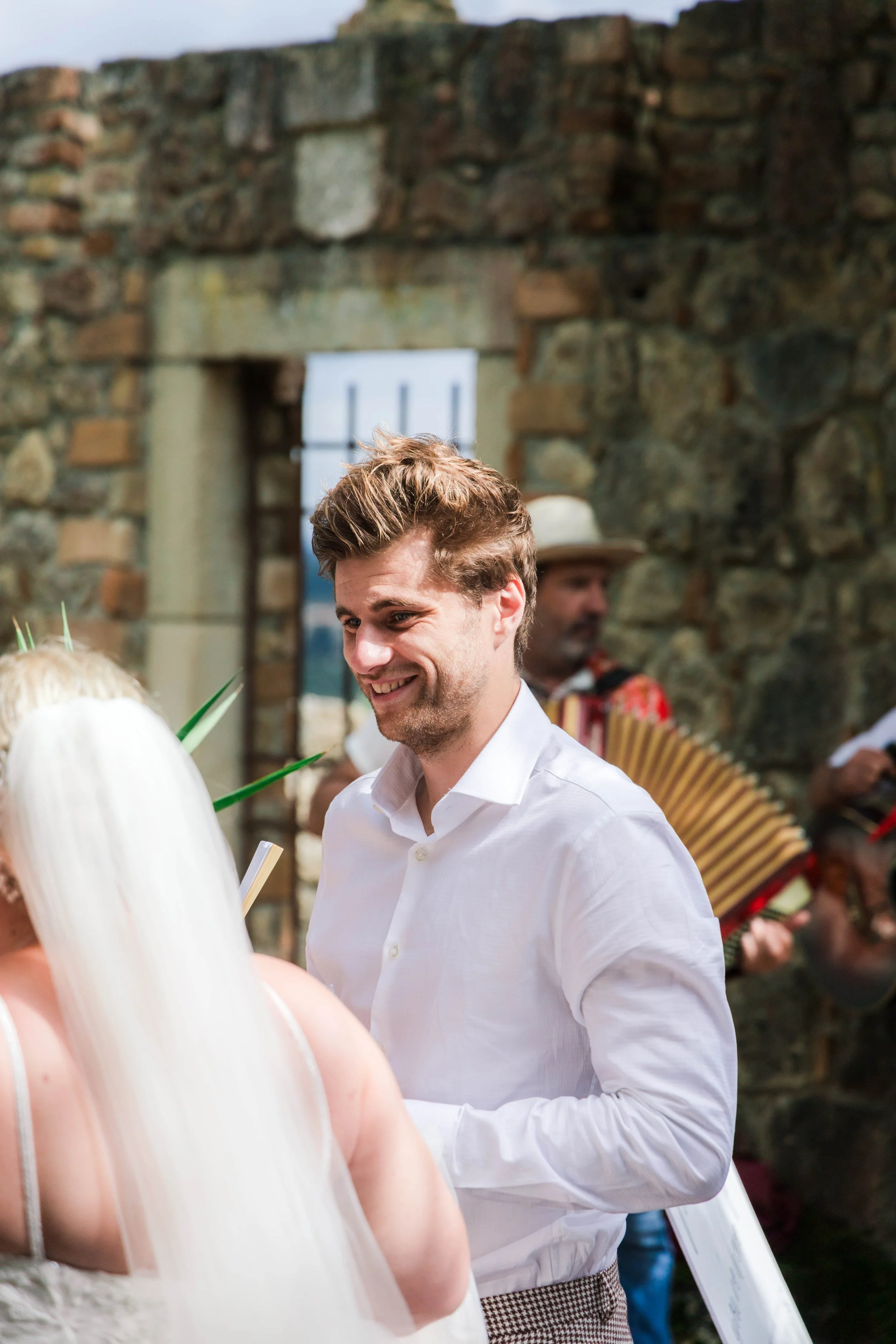 Young man in a white shirt smiling. Stone wall and other people, one playing an accordion, in the background.