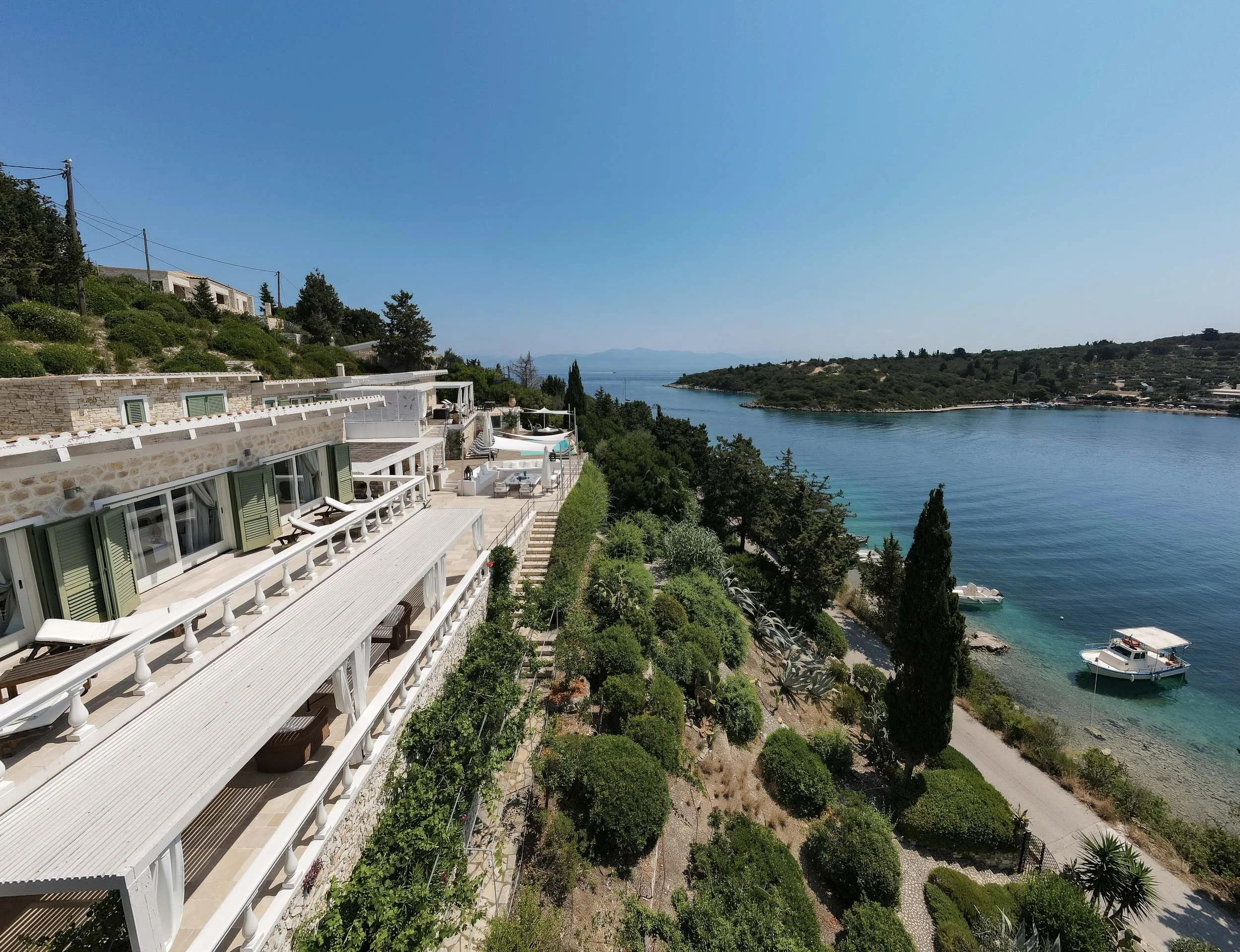 A seaside view of a stone building with white balconies and green shutters, overlooking a lush hillside and calm blue water with boats, under a clear blue sky.