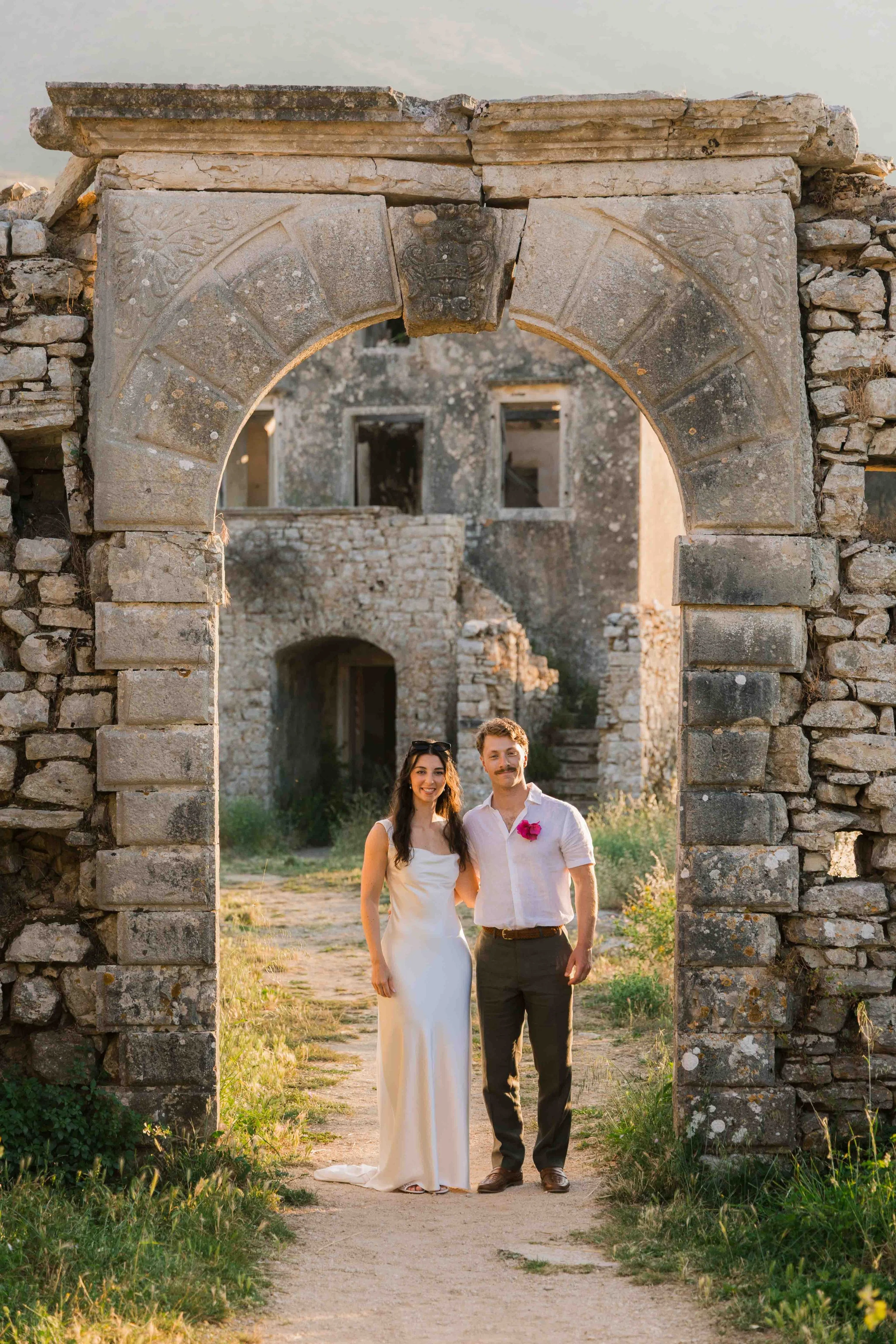 A couple standing hand in hand in front of an ancient stone archway with a historic stone building in the background, during sunset.