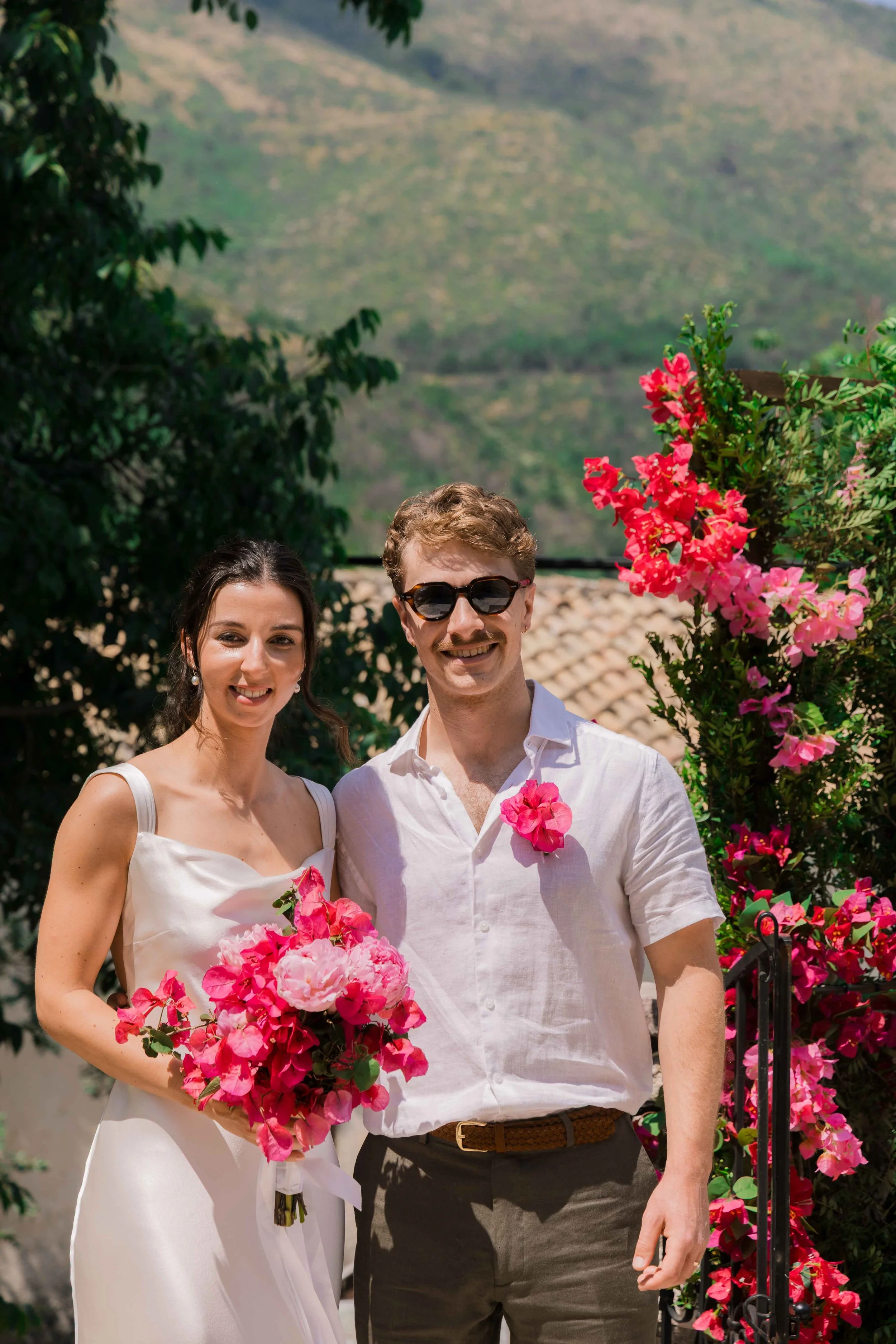 A bride and groom stand together outdoors during a wedding, smiling. The bride wears a white dress and holds a bouquet of pink and red flowers. The groom wears sunglasses, a white shirt, and has a pink flower pinned to his shirt. Colorful flowers and