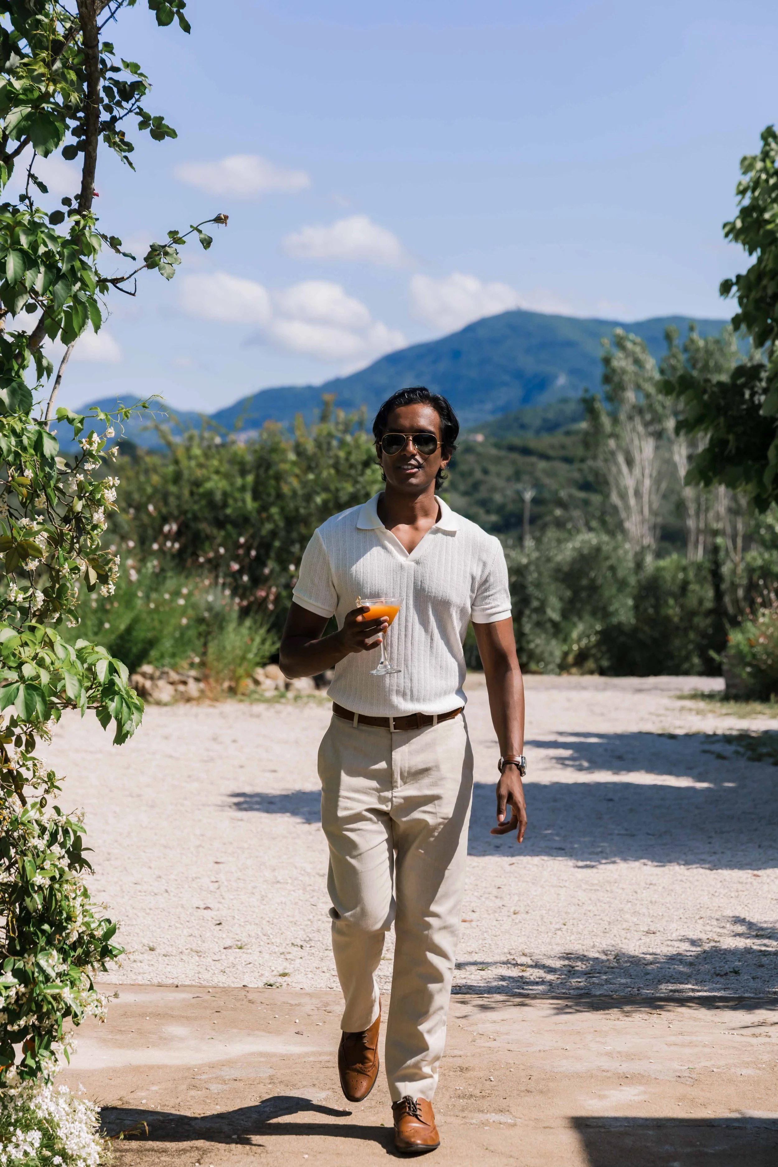 A man walking outdoors on a sunny day, holding a cocktail, with mountains and a blue sky in the background.