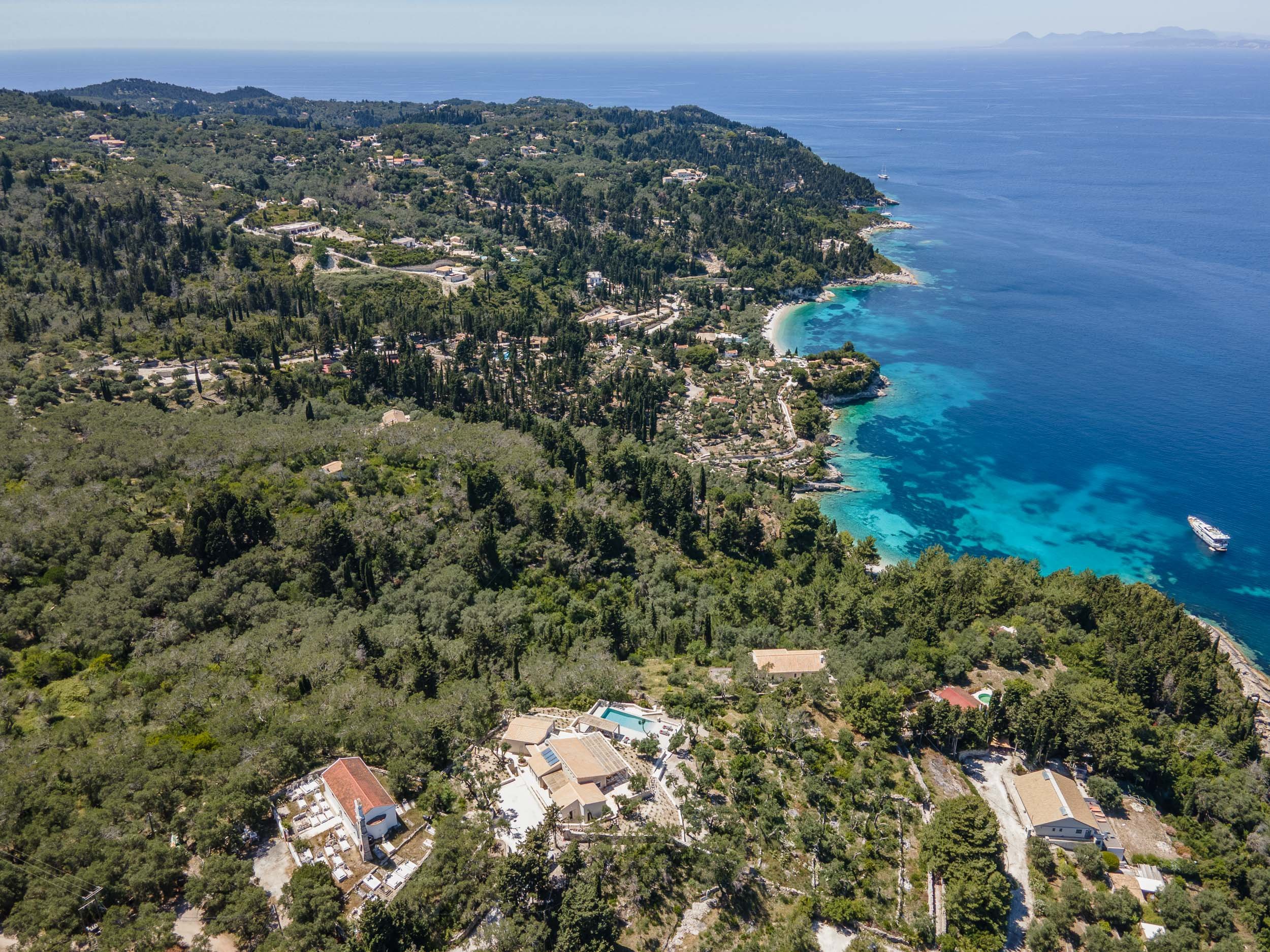 Aerial view of a coastal landscape with dense green trees, houses, and a clear blue ocean with visible underwater features, boats, and a sandy shoreline.