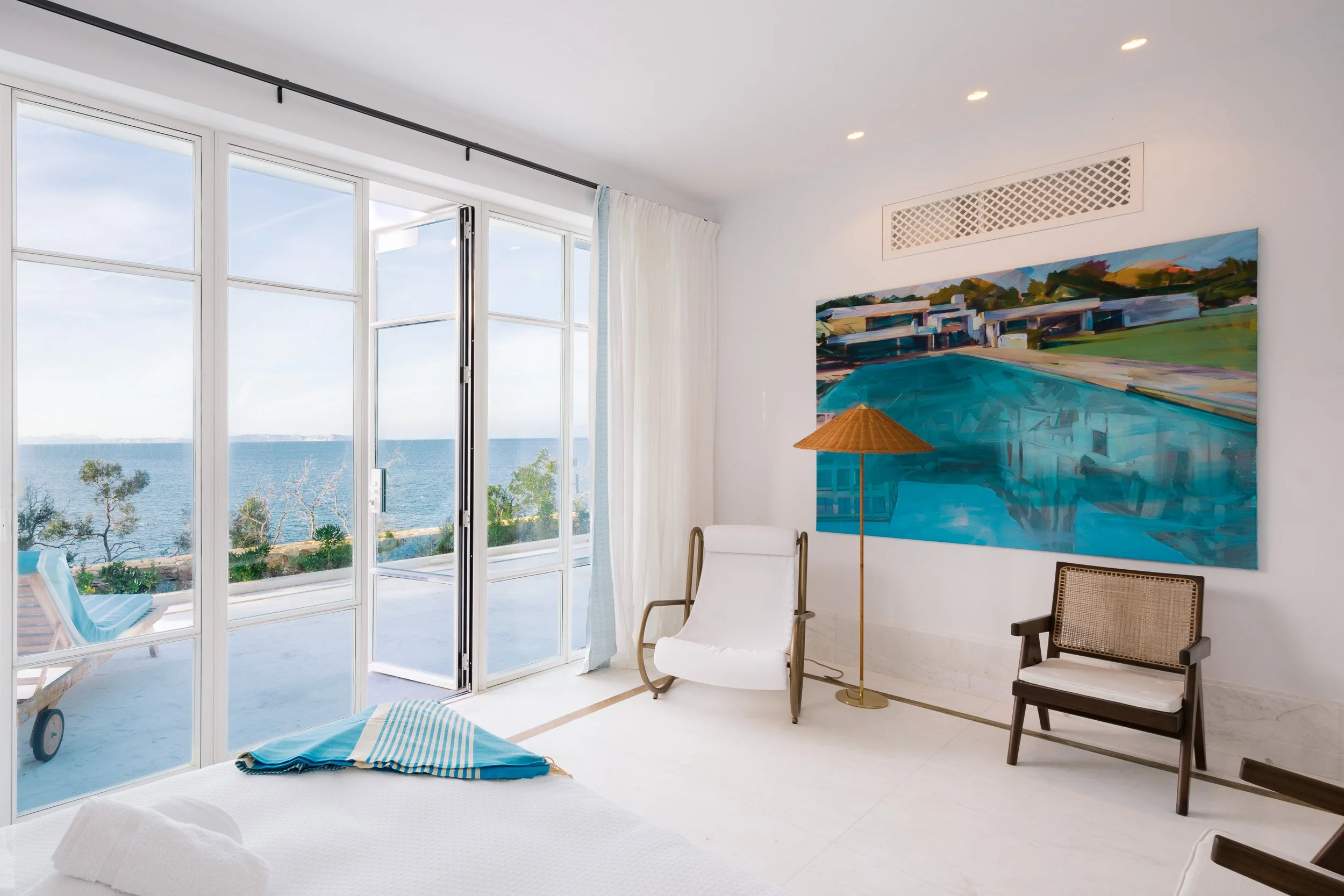 A bright hotel room with large glass doors offering a view of the ocean, featuring sea, trees, and outdoor lounge chairs. The room has a white bed, a white chair, a colorful abstract painting, a wicker basket chair, and a floor lamp.