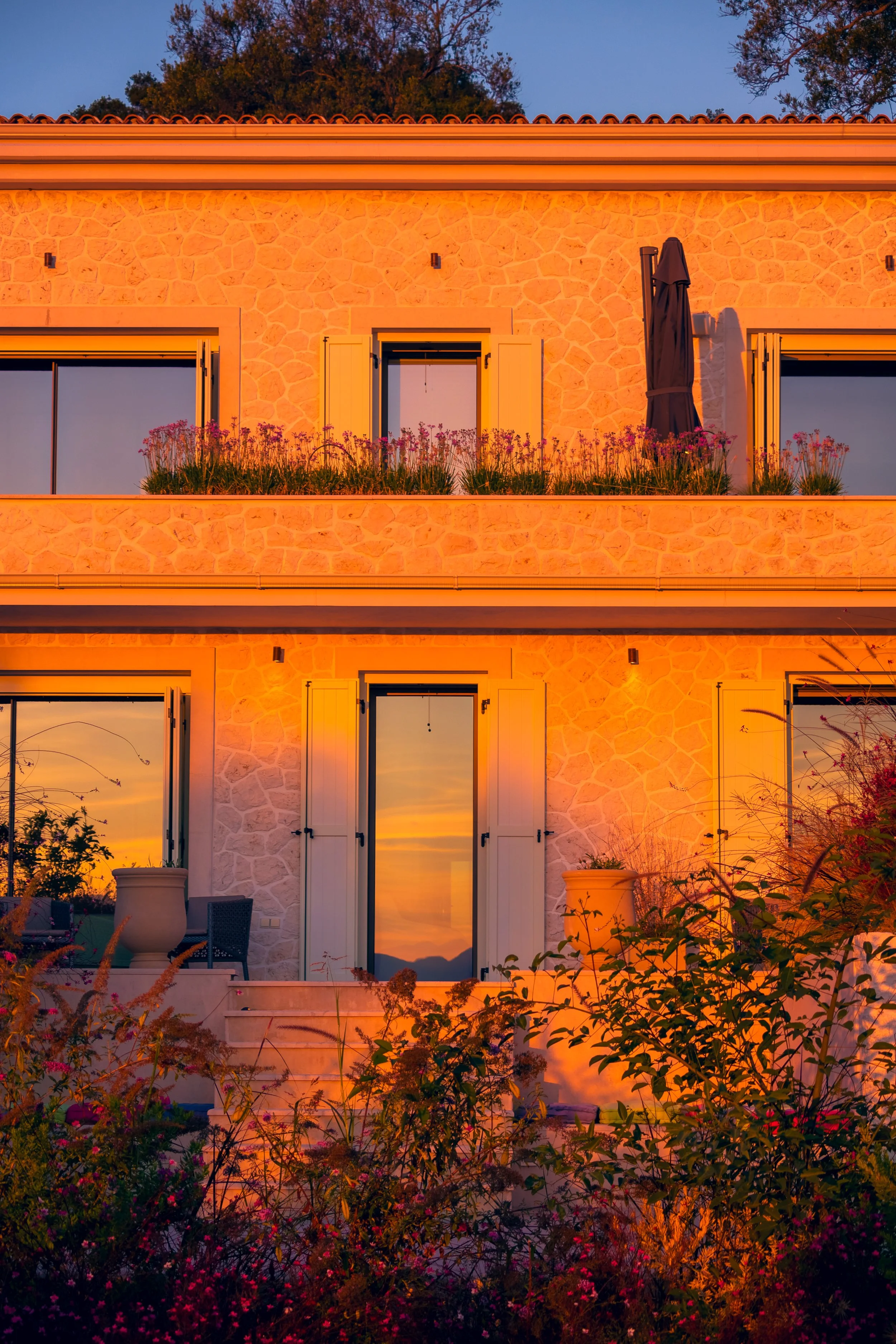 A two-story house at sunset with stone exterior walls, open windows with shutters, and a balcony with potted plants, reflecting the colorful sky in the glass doors and windows.