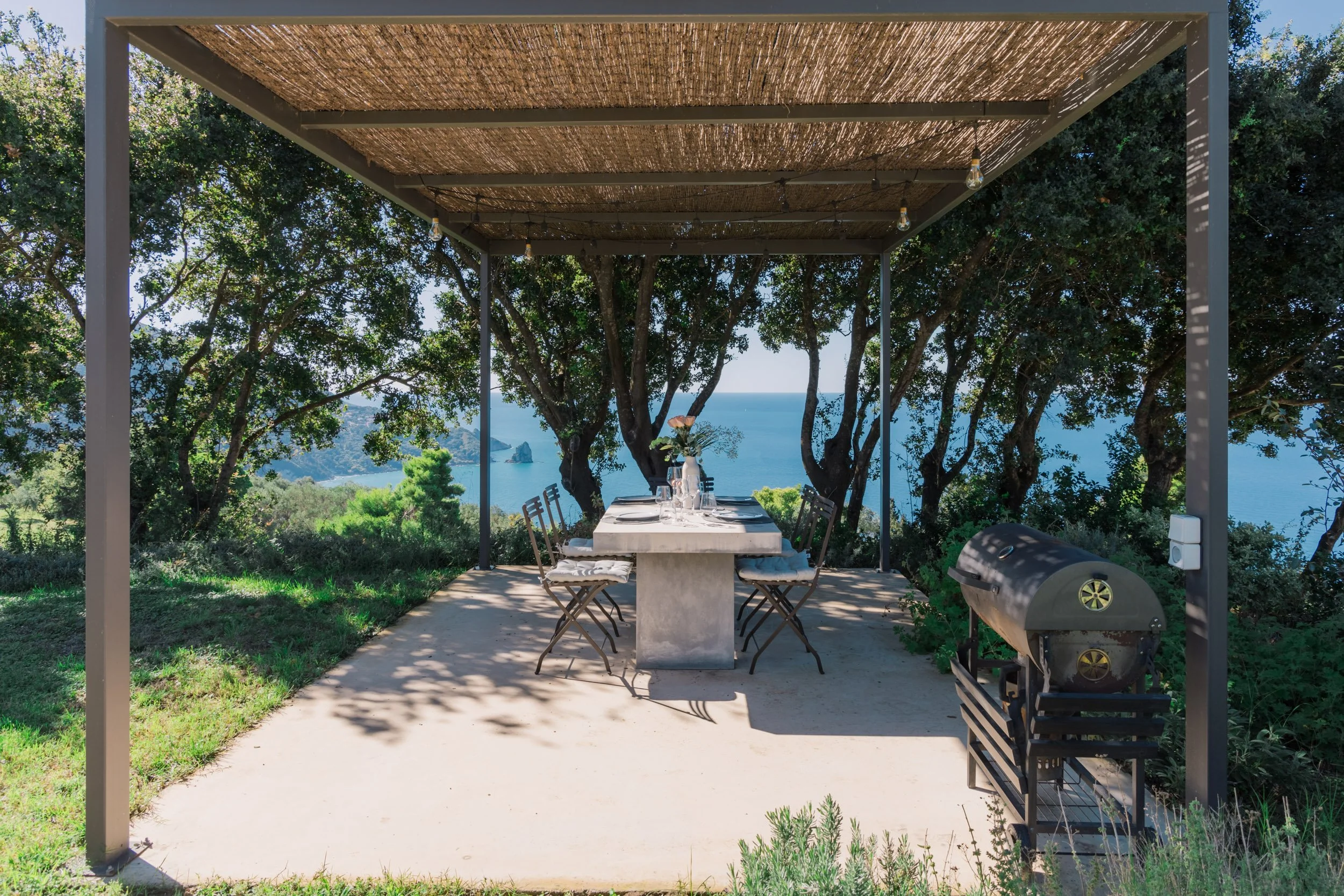 Outdoor dining area under a pergola with a view of trees and the ocean in the background, featuring a dining table with chairs, a vase with flowers, and a barbecue grill.