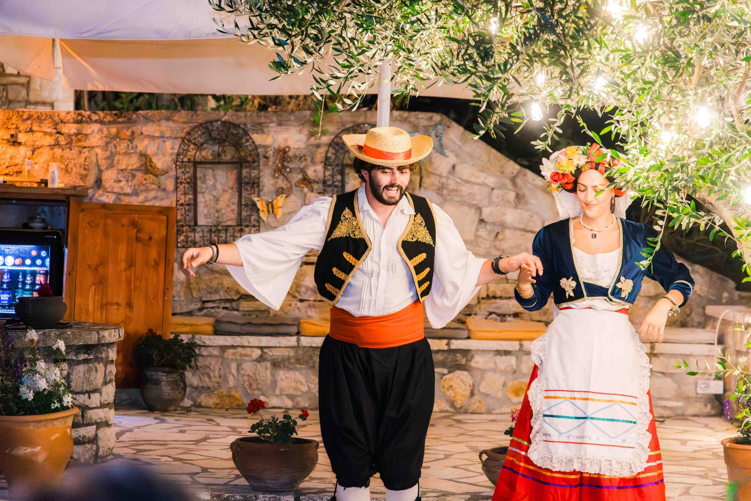 A man and woman dressed in traditional folk costumes, dancing outdoors under string lights in front of a stone wall and greenery.
