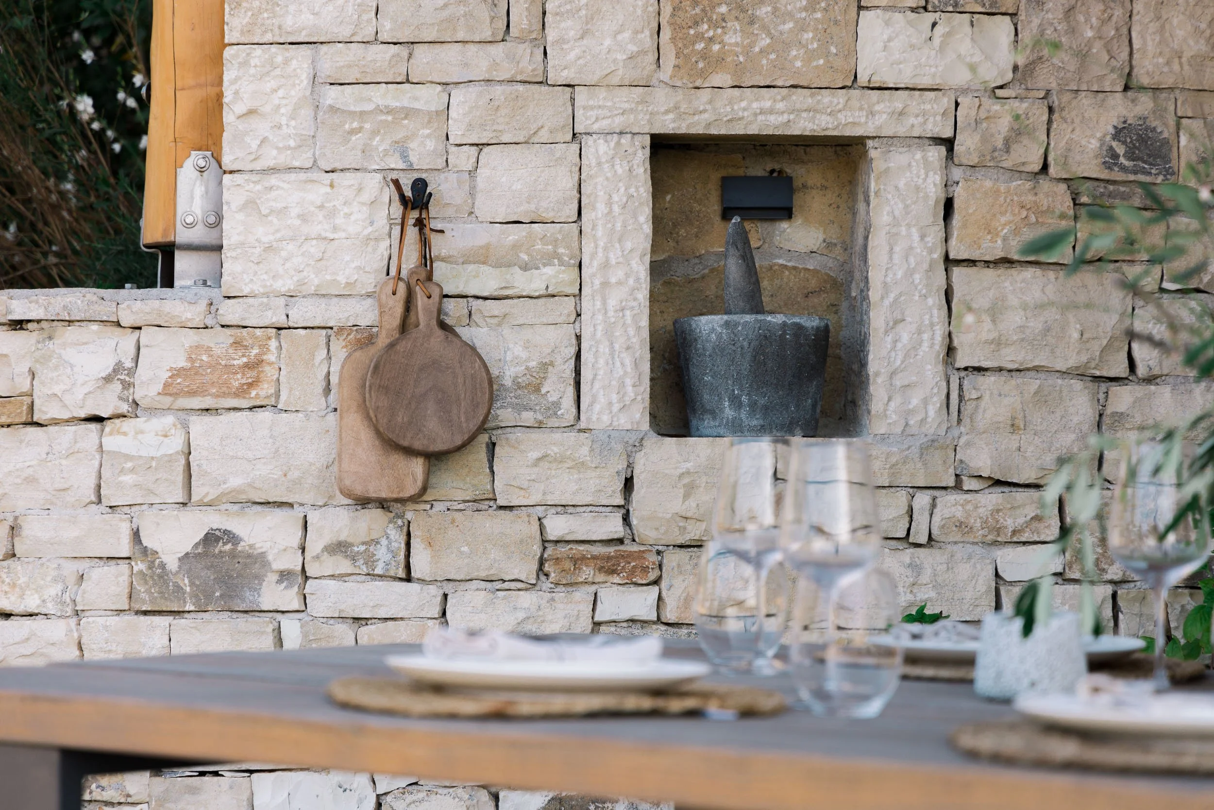 Outdoor dining table with glassware and dishes in front of a beige stone wall. A small water fountain with a stone pestle and bowl is in a niche in the wall, with hanging wooden cutting boards nearby.