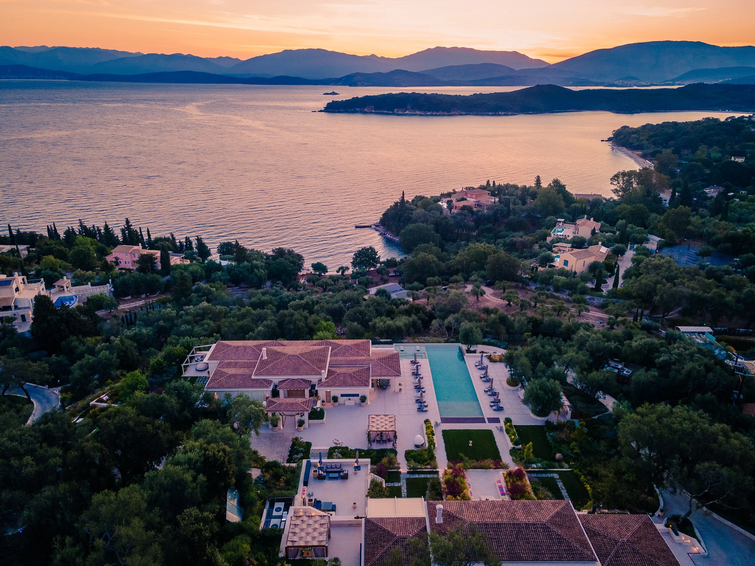 Aerial view of a lakeside residential area during sunset with houses, a swimming pool, and lush greenery.