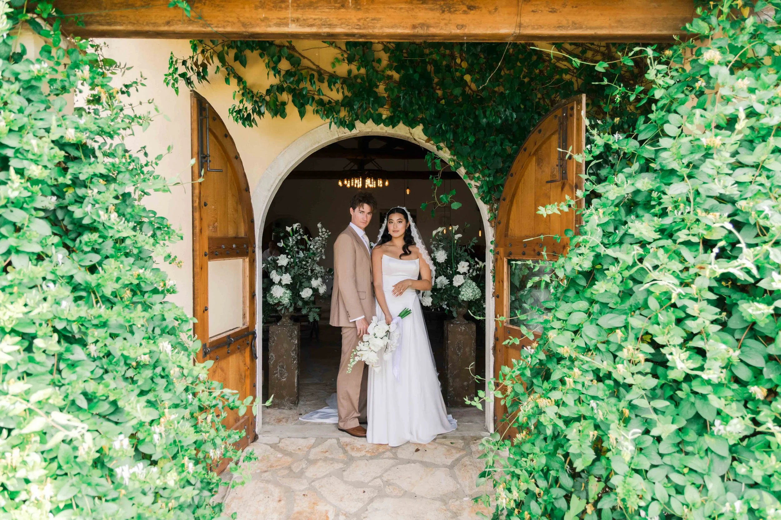 A bride and groom stand at the entrance of a wedding venue with open wooden doors, surrounded by lush green foliage. The bride wears a white wedding gown with a veil and holds a bouquet of white flowers, while the groom wears a beige suit and white s