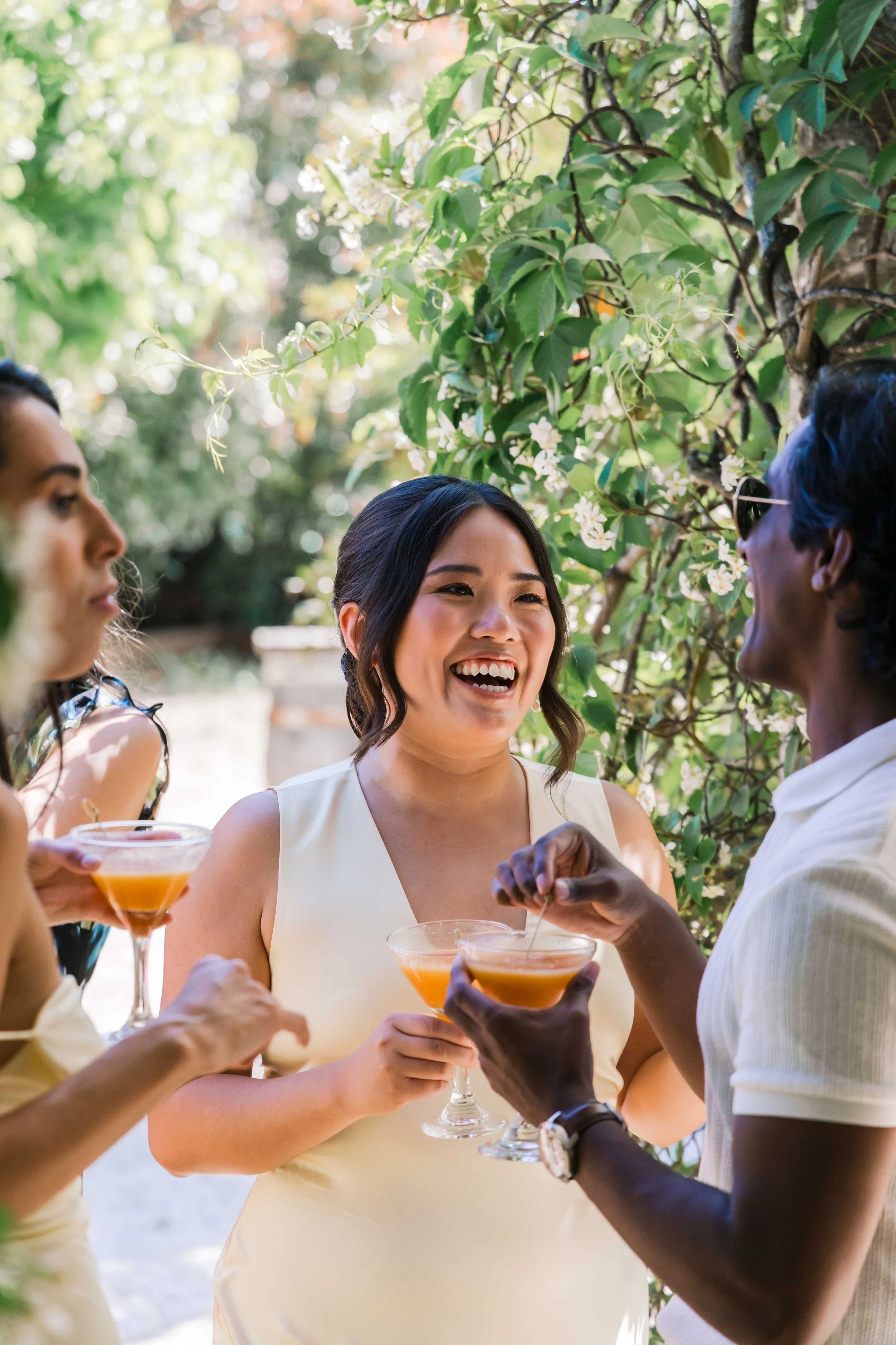 Four women socializing outdoors, holding cocktails, with greenery and flowering vine in the background, one woman smiling and talking.