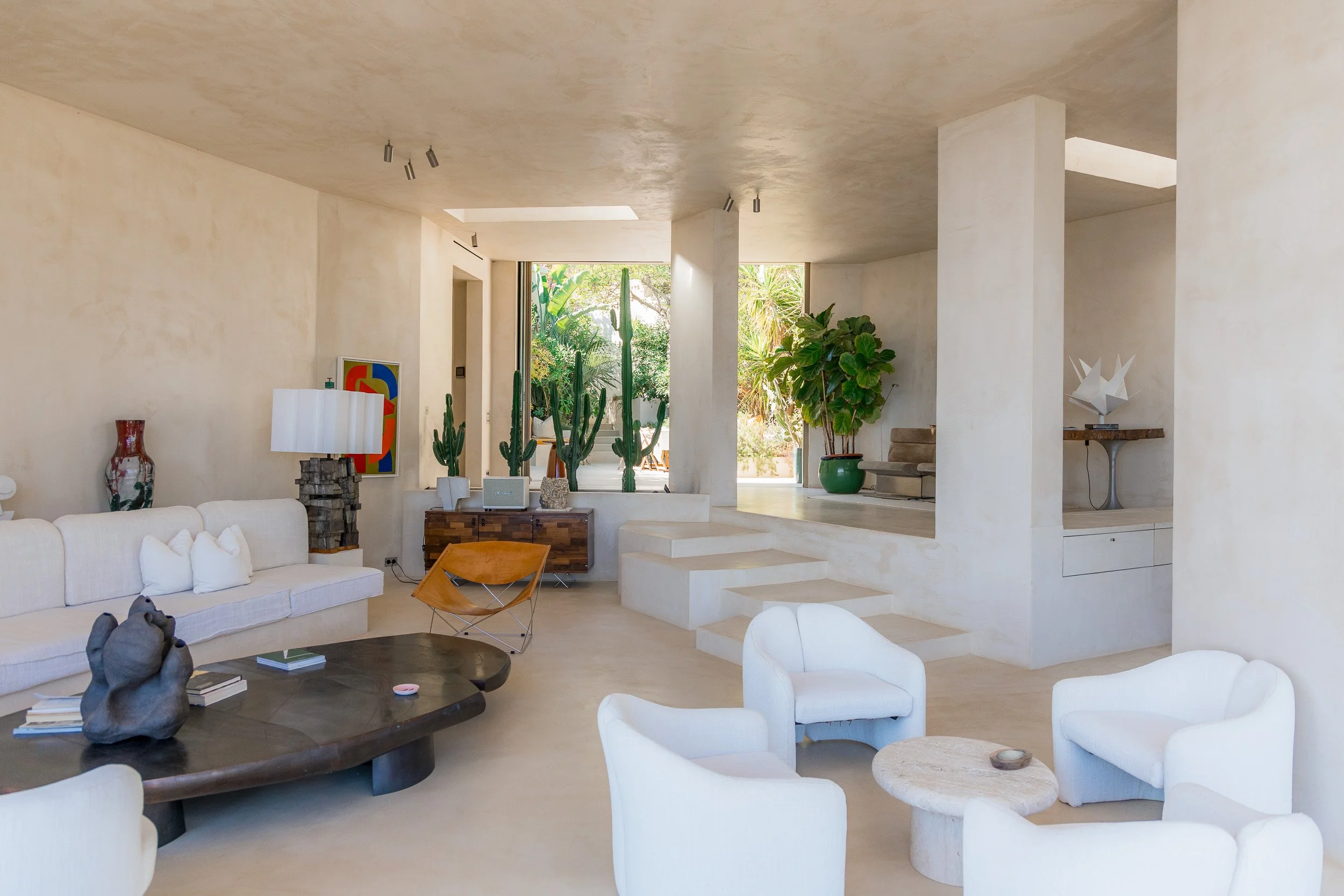 Bright, minimalist living room with white furniture, large green plants, and natural light from large windows.