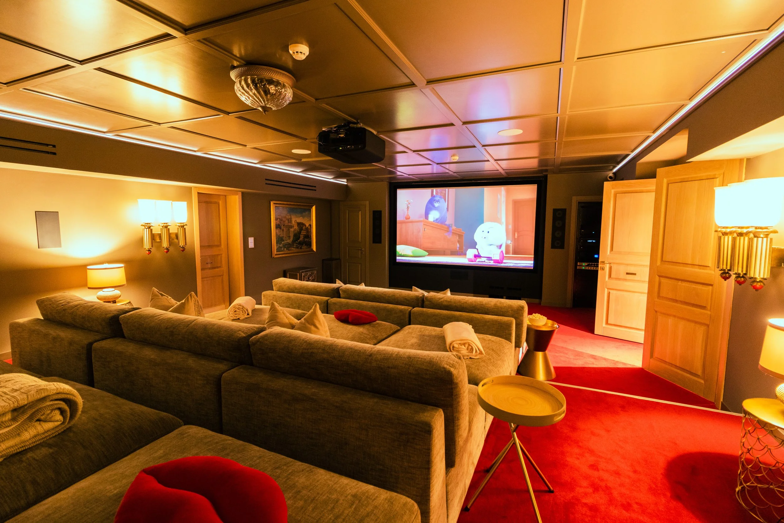 A cozy home theater room with beige couches, a large screen displaying animated characters, wooden ceiling panels, and warm lighting.