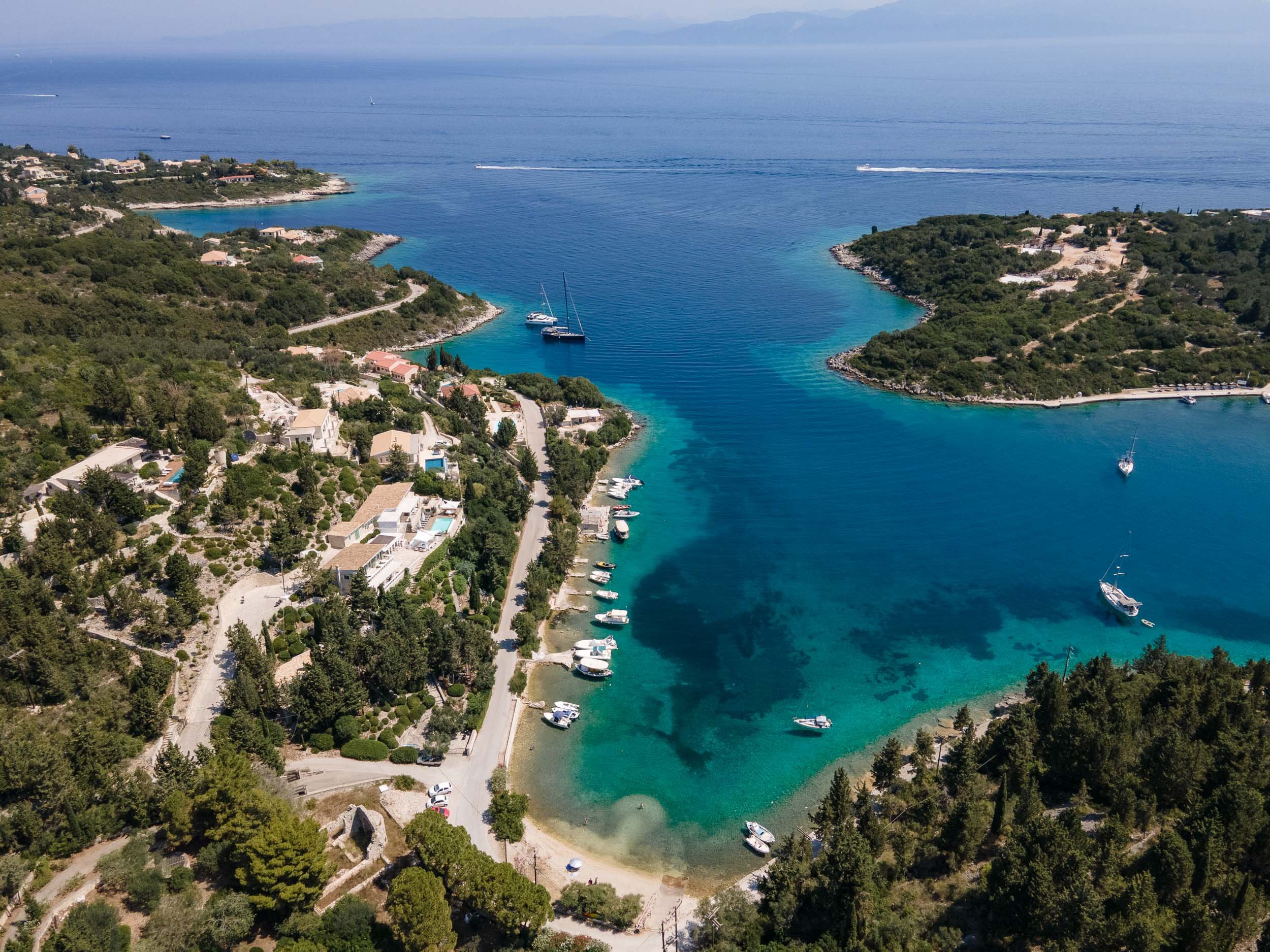 Aerial view of a coastal area with clear blue water, boats docked near the shore, residential houses surrounded by greenery, and lush hills in the background.