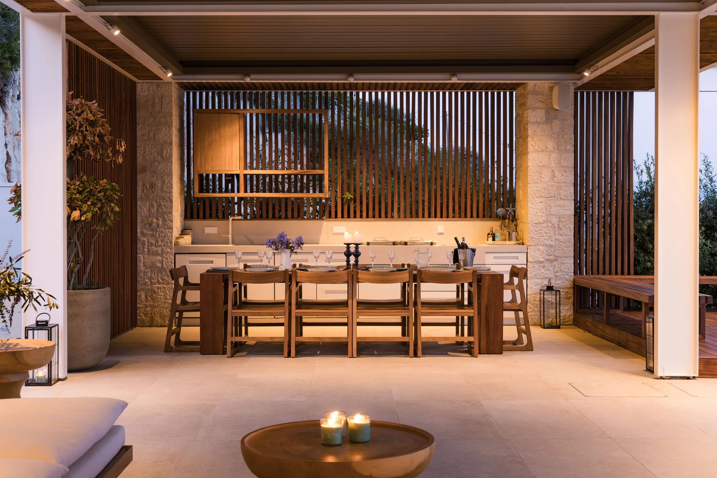 Outdoor dining area with a large wooden table, chairs, a centerpiece with purple flowers, and candles, under a covered patio with wooden and stone accents.