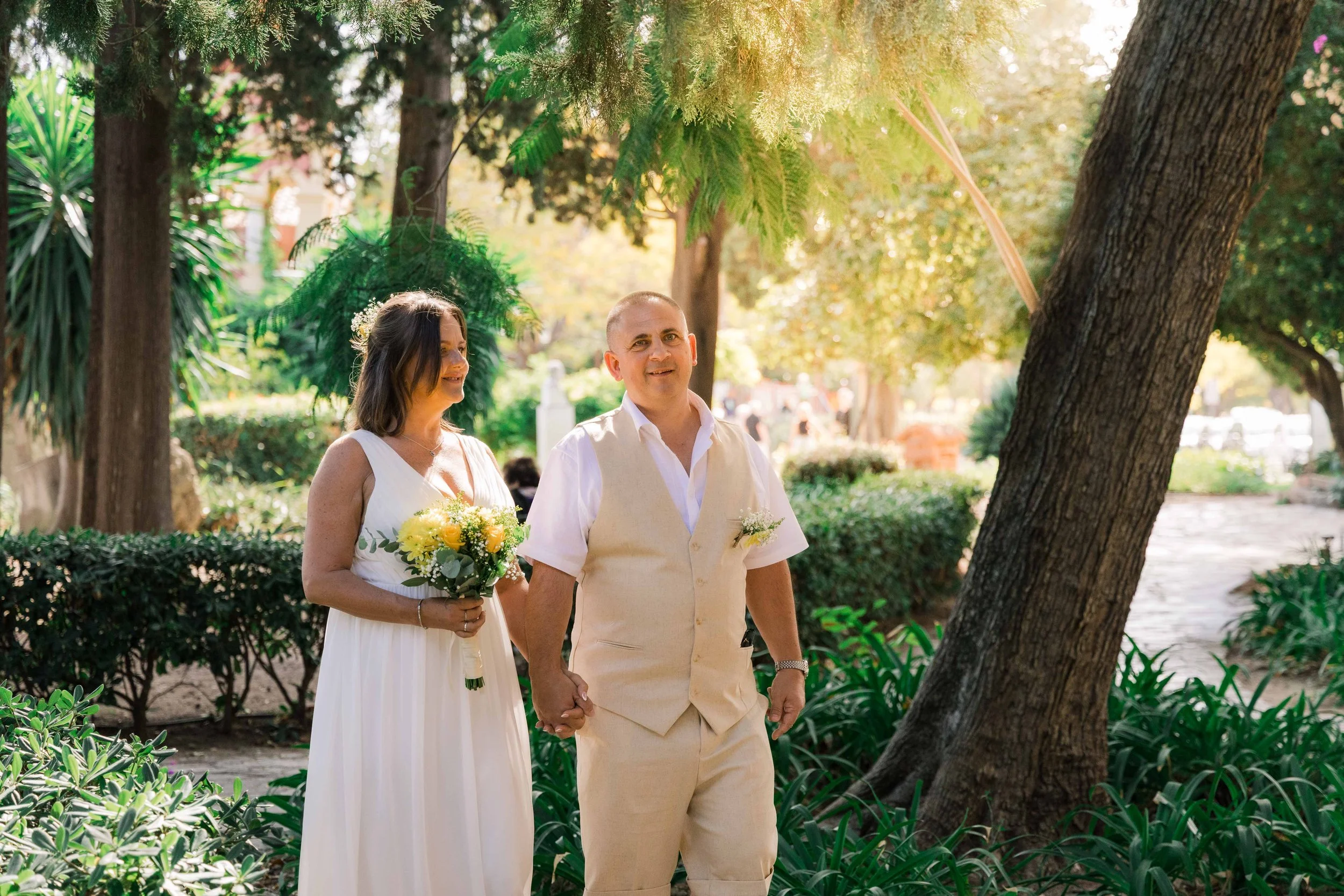 A couple stands outdoors in a park, holding hands, with the woman holding a bouquet of yellow and white flowers. They are surrounded by lush greenery and trees, with sunlight filtering through the leaves.