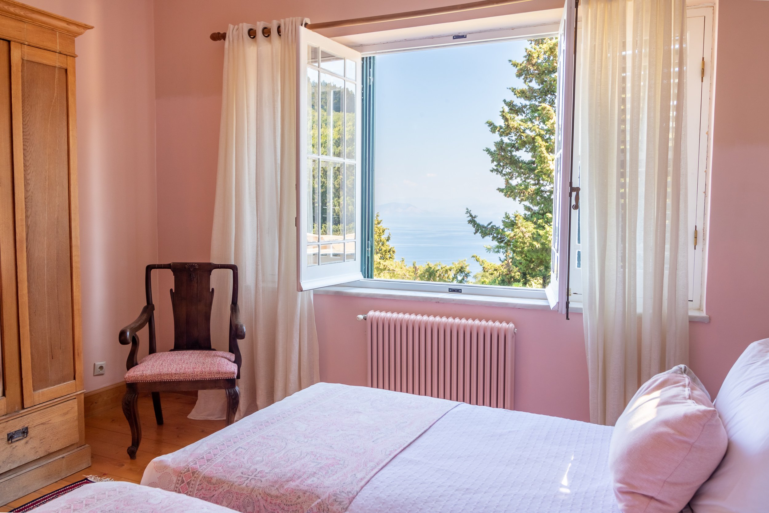 A bedroom with an open window showing a view of trees and water outside. The room has pink walls, white curtains, a wooden wardrobe, a wooden chair with pink upholstery, and a bed with white bedding.