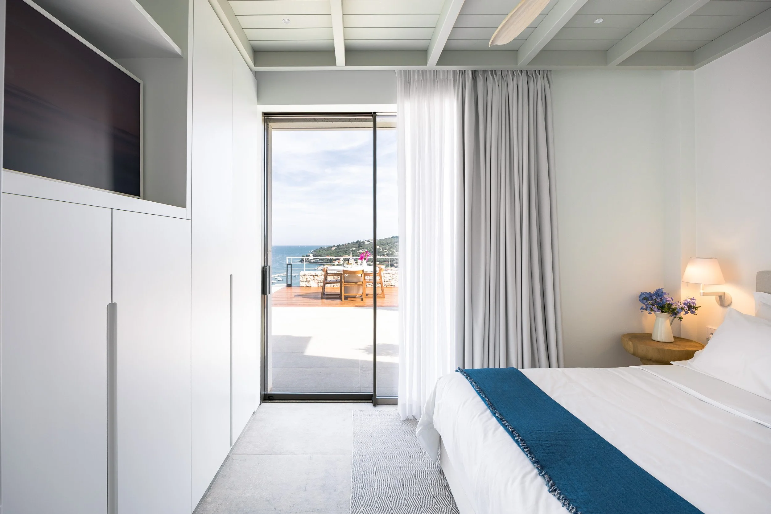 A bright hotel room with a bed, a nightstand with a flower vase, and a sliding glass door leading to a balcony overlooking the ocean and a nearby hill.