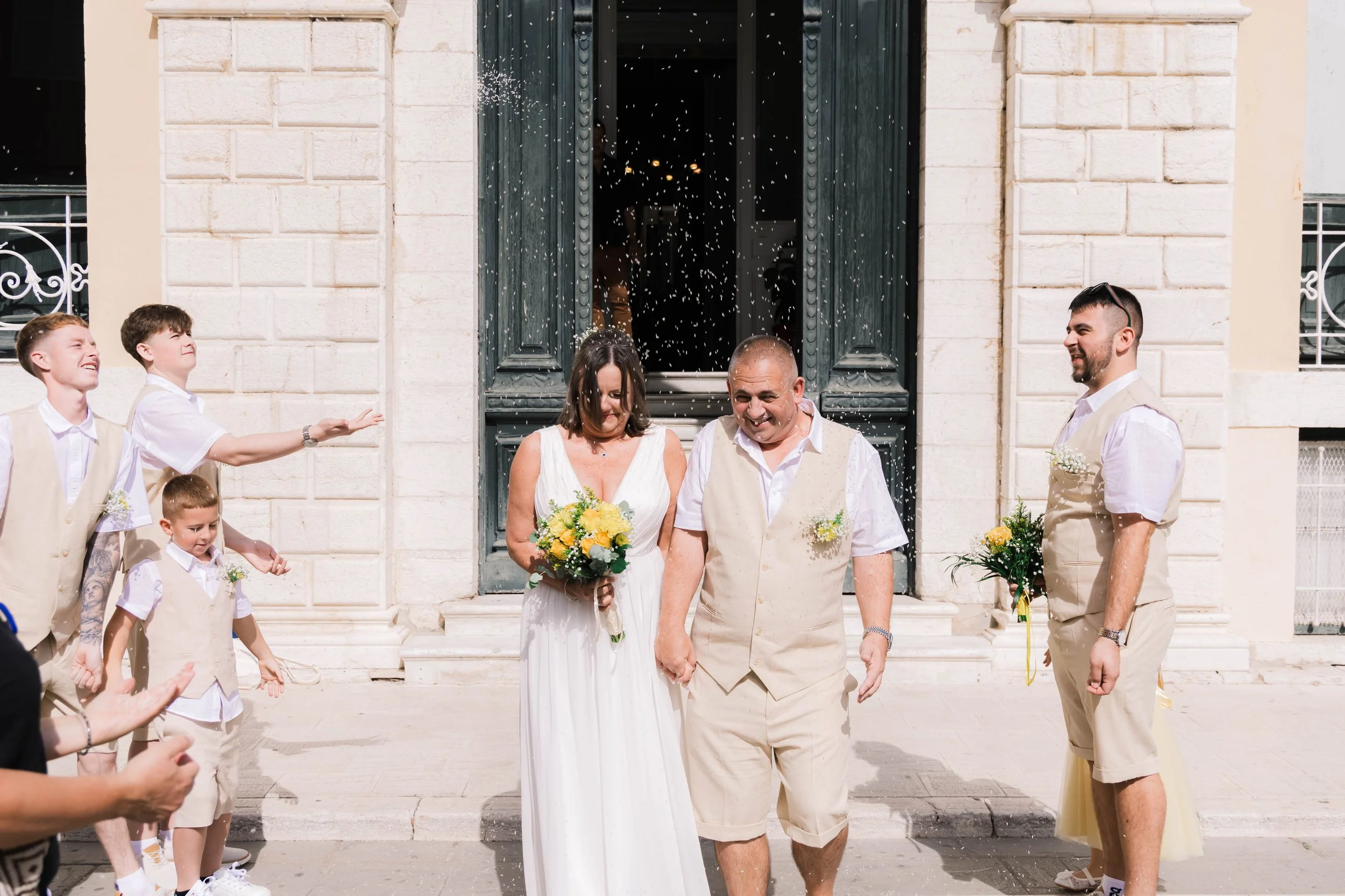 A couple is walking hand in hand after a wedding ceremony outside a building with large doors. They are surrounded by children and other guests, some of whom are throwing rice or confetti, celebrating as they rejoice.