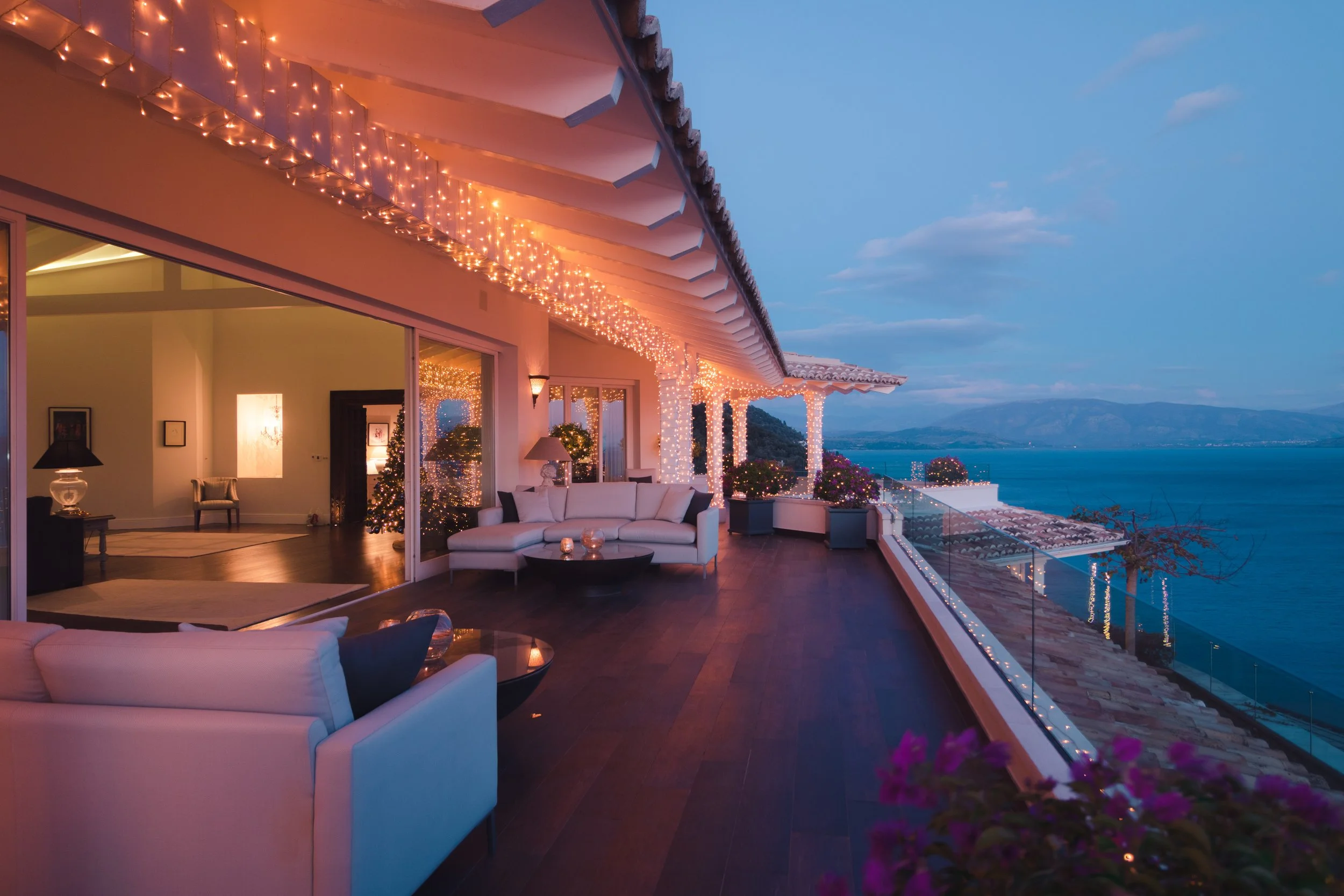 A spacious balcony decorated with string lights, overlooking a body of water with mountains in the distance, with outdoor seating, potted plants, and a view into a well-lit interior living space.