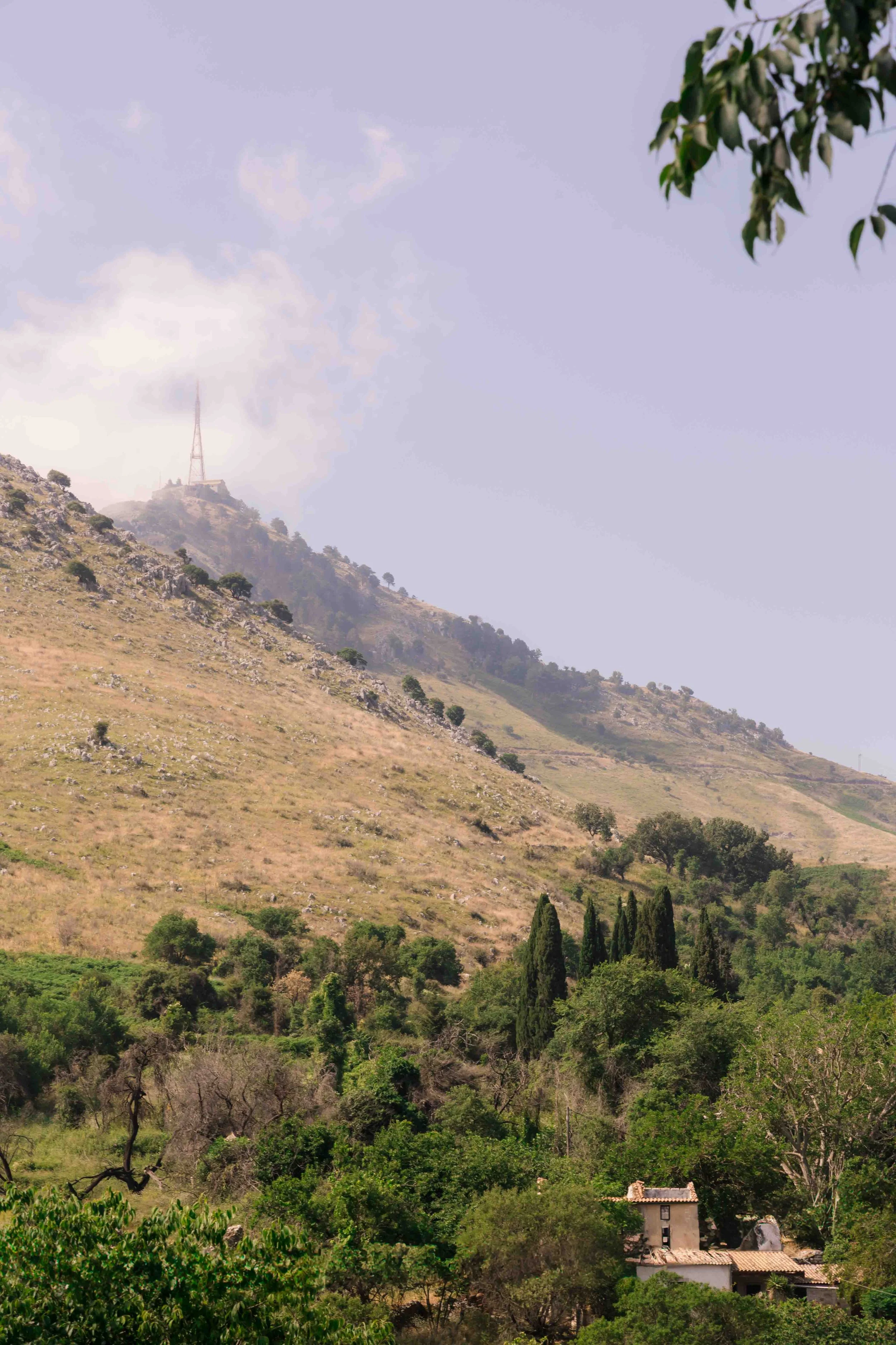 Scenic view of a hilly landscape with a small house at the bottom, surrounded by trees, and a mountain in the background with a communication tower near the top. The sky is partly cloudy.