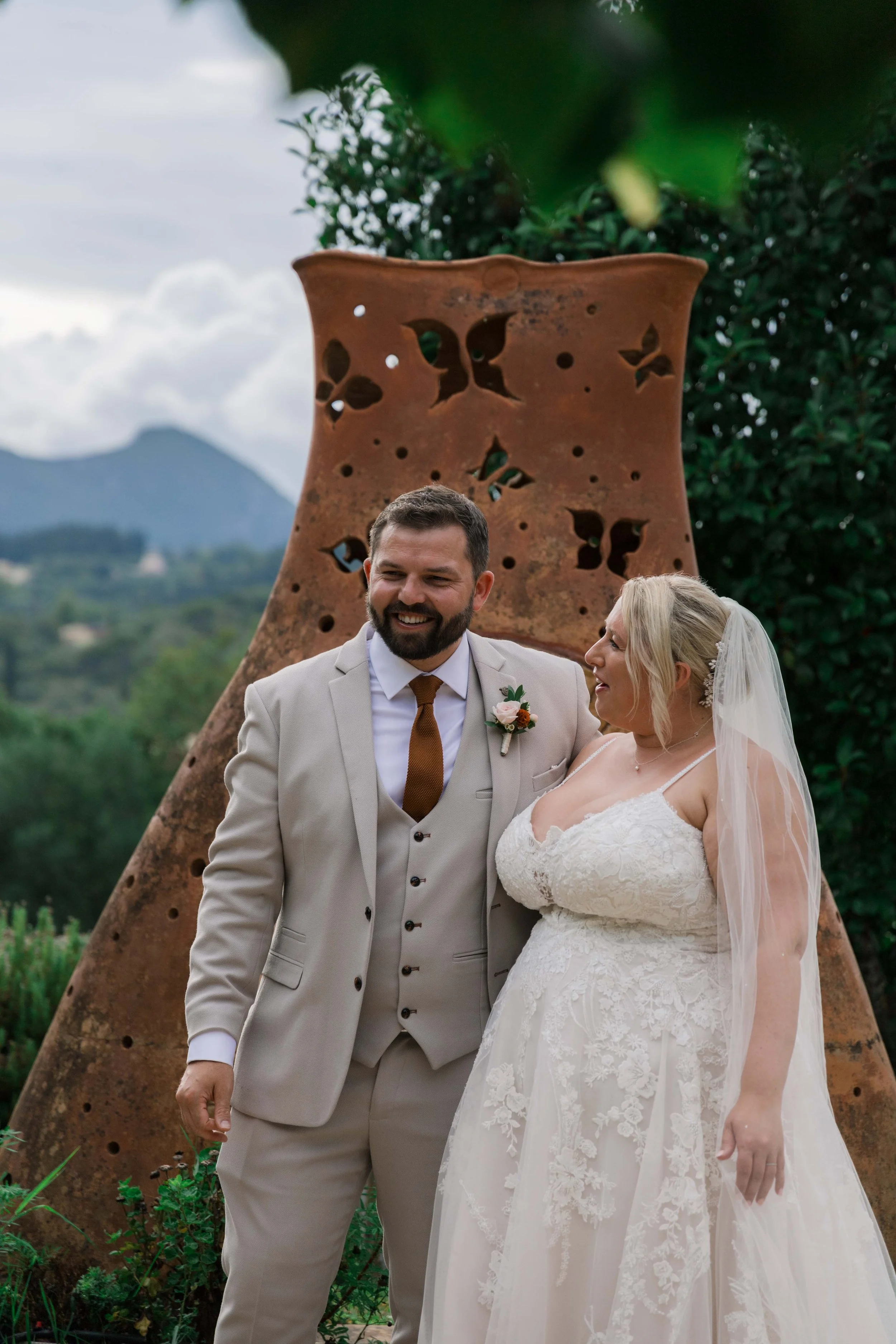 A bride and groom are smiling together outdoors during their wedding, with a large, decorative, butterfly-shaped metal sculpture in the background and mountains in the distance.