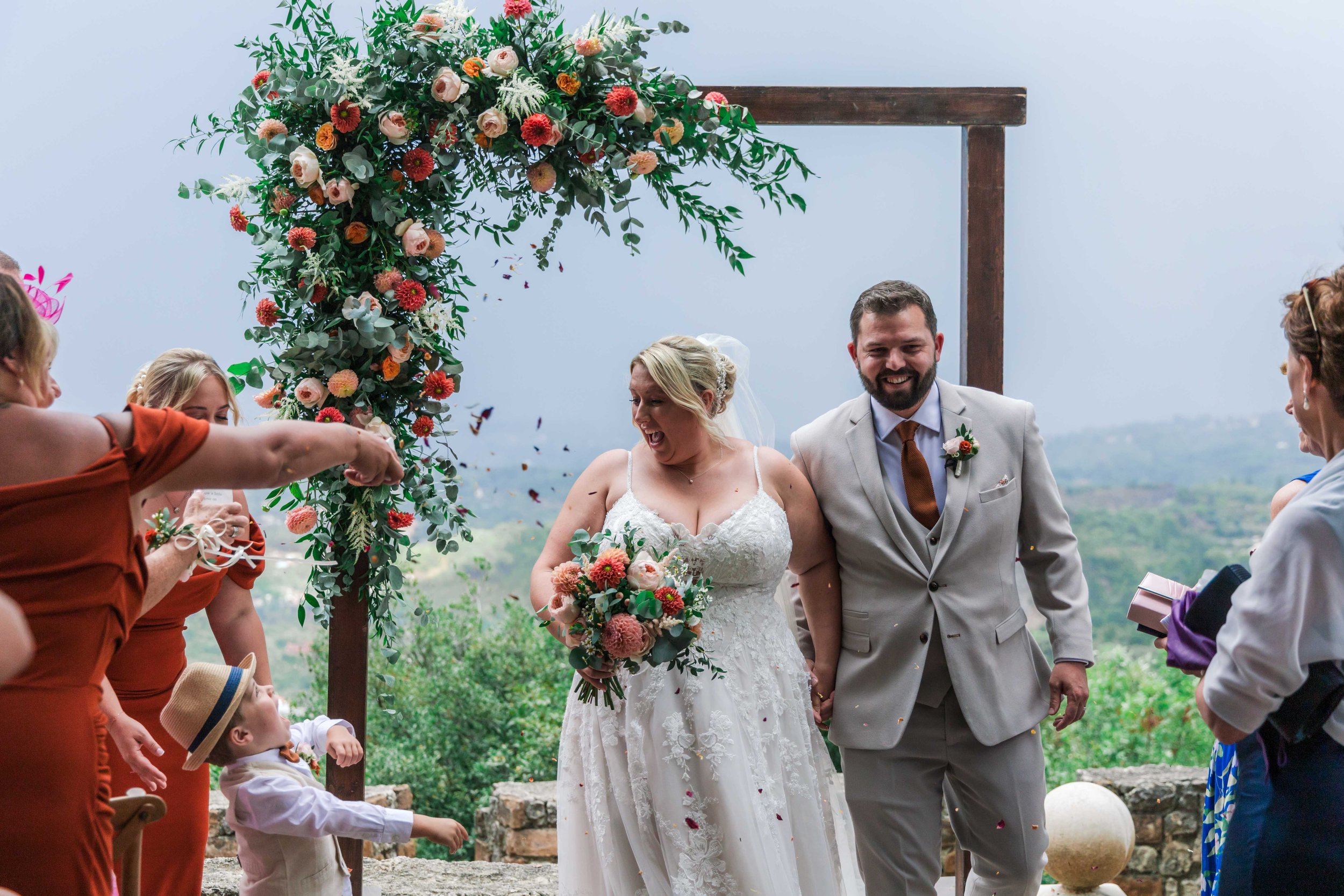 Bride and groom holding hands under a floral arch at an outdoor wedding ceremony, surrounded by friends and family, with a scenic landscape in the background.