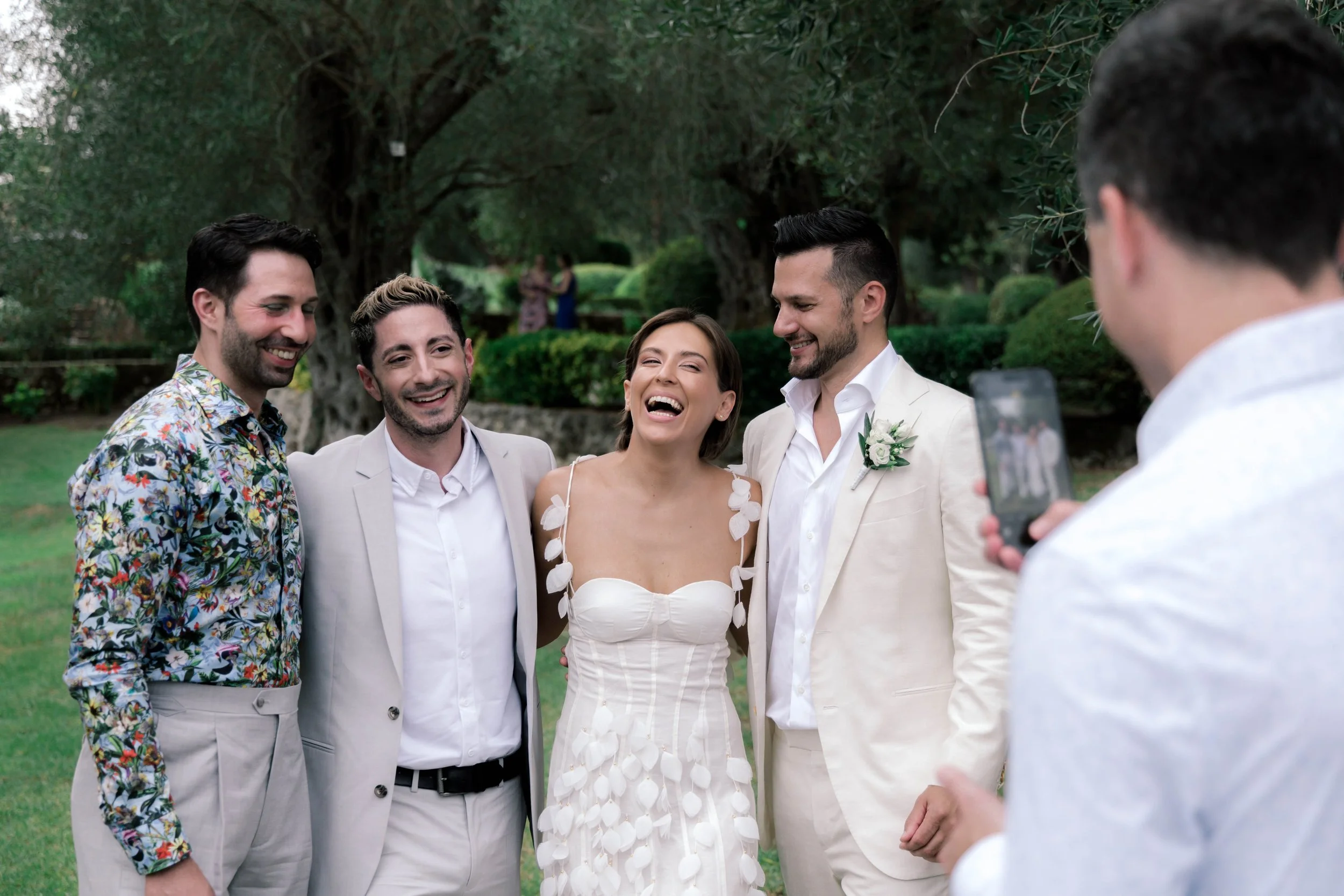 Group of five friends taking a photo together at an outdoor celebration, with greenery in the background.