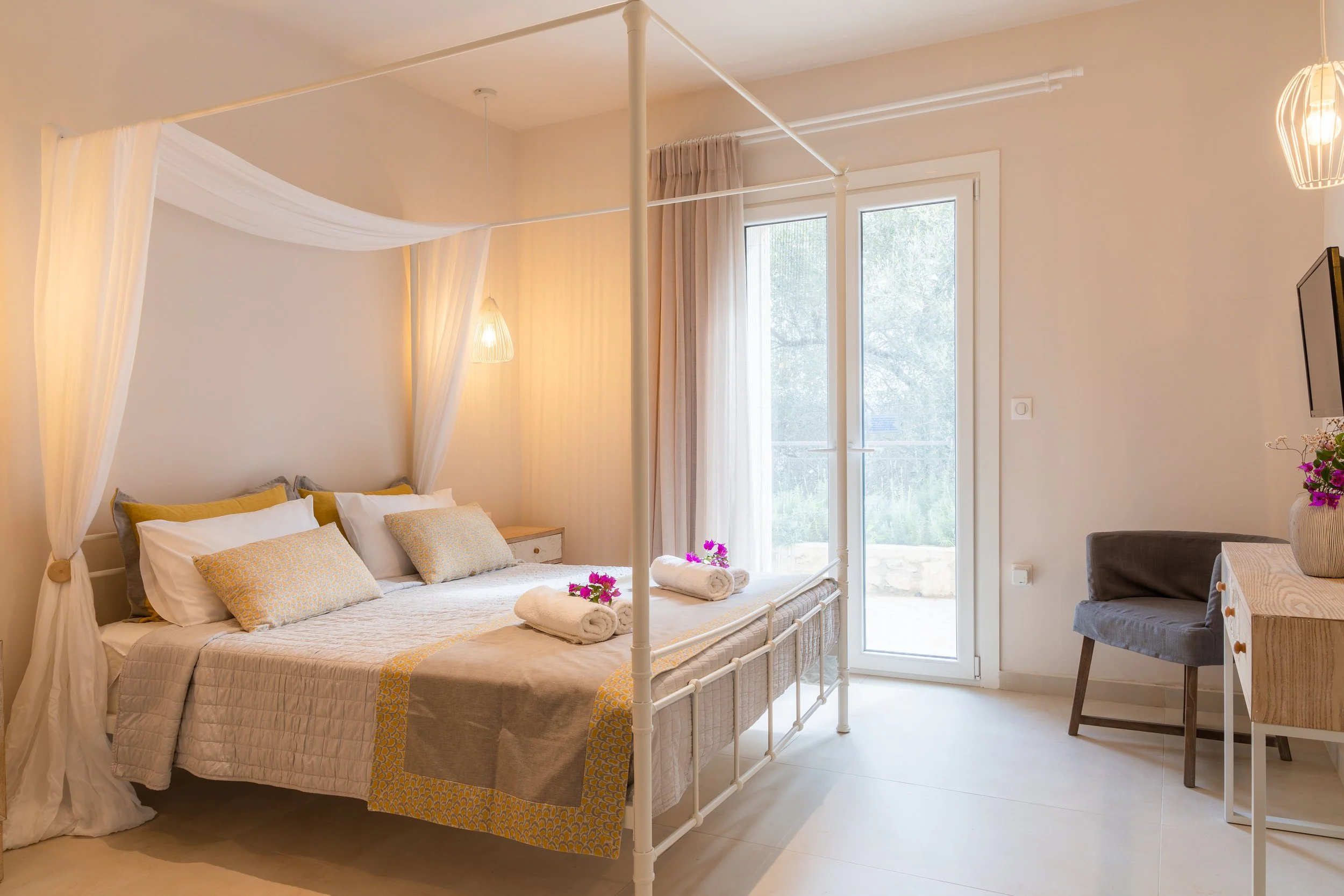 A bright, modern bedroom with a white four-poster canopy bed, decorated with beige and yellow accents, accompanied by rolled towels and pink flowers. There is a large window with sheer curtains allowing natural light, and furniture including a gray c