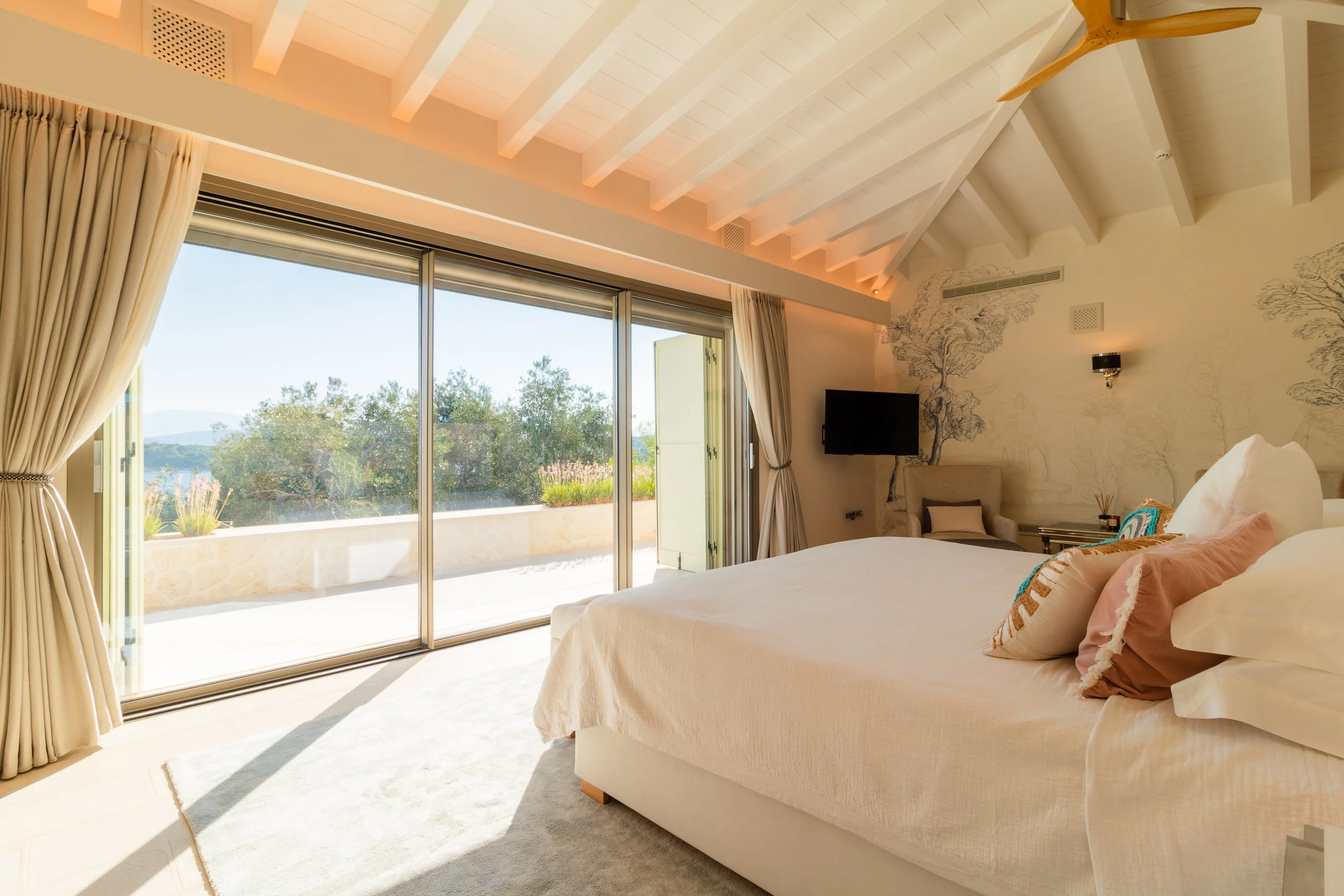 Bright hotel bedroom with a large bed, beige and pink pillows, sliding glass doors leading to a balcony with outdoor plants, and a scenic view of trees and mountains outside.