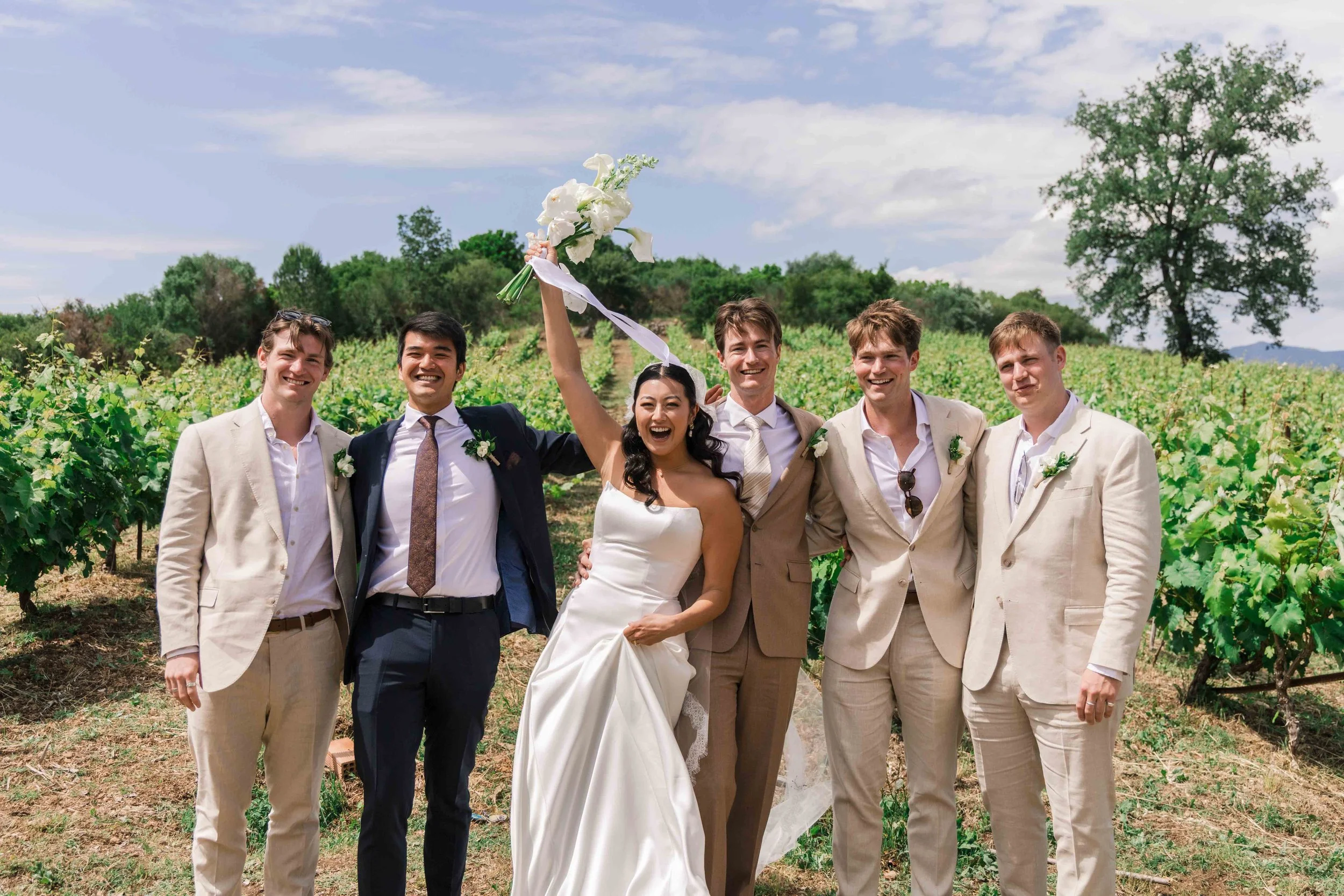 A bride and five groomsmen celebrating outdoors in a vineyard, with the bride holding a bouquet, all smiling and dressed in wedding attire.