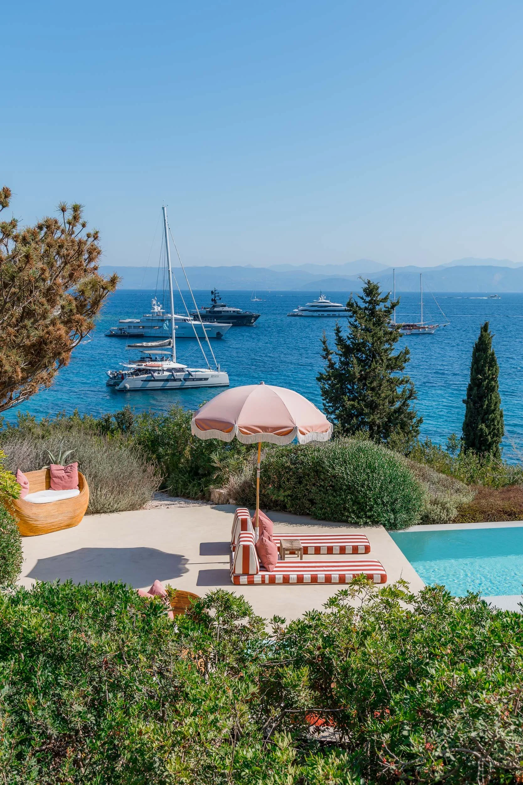 A scenic view of a coastline with boats anchored offshore, a swimming pool with striped lounge chairs and a pink striped umbrella in the foreground, surrounded by green bushes and trees.