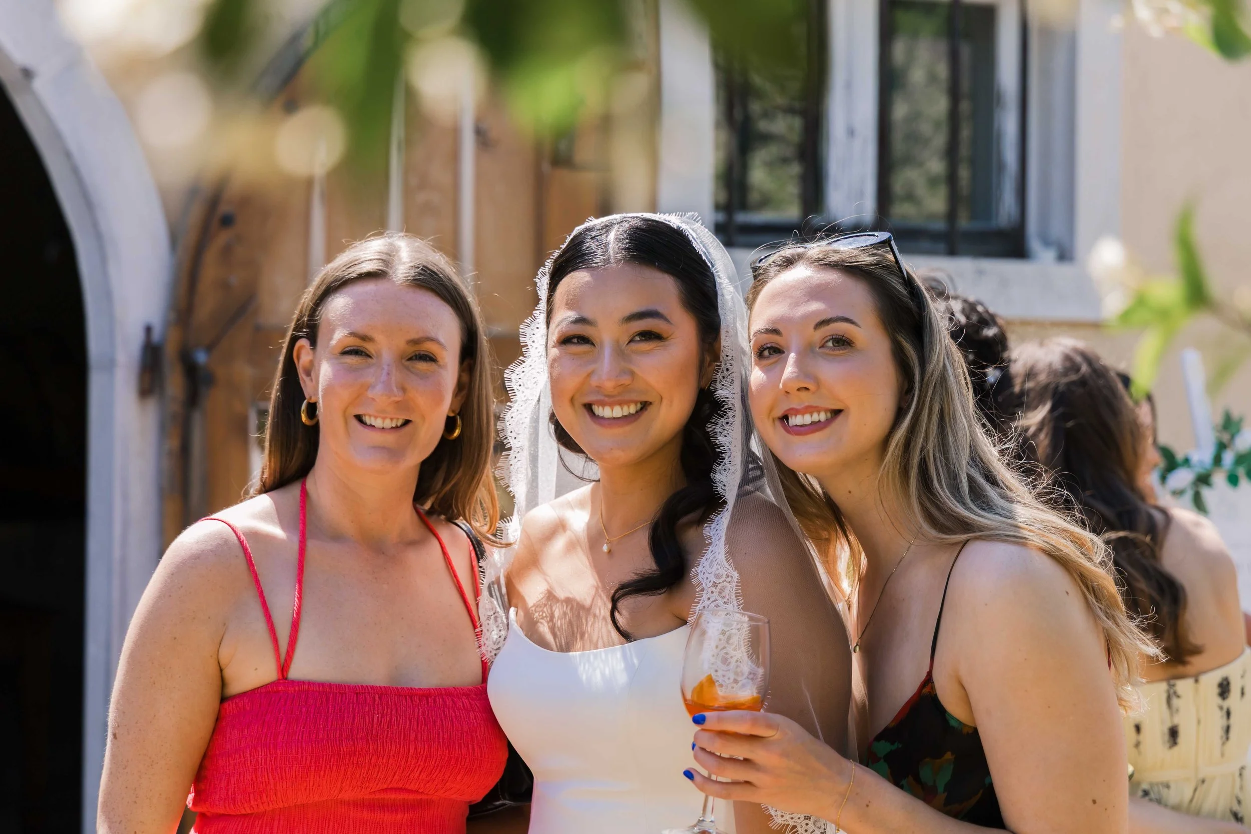 Three women smiling outdoors, one wearing a wedding veil and holding a glass of wine, others in summer dresses, at a social gathering or celebration.