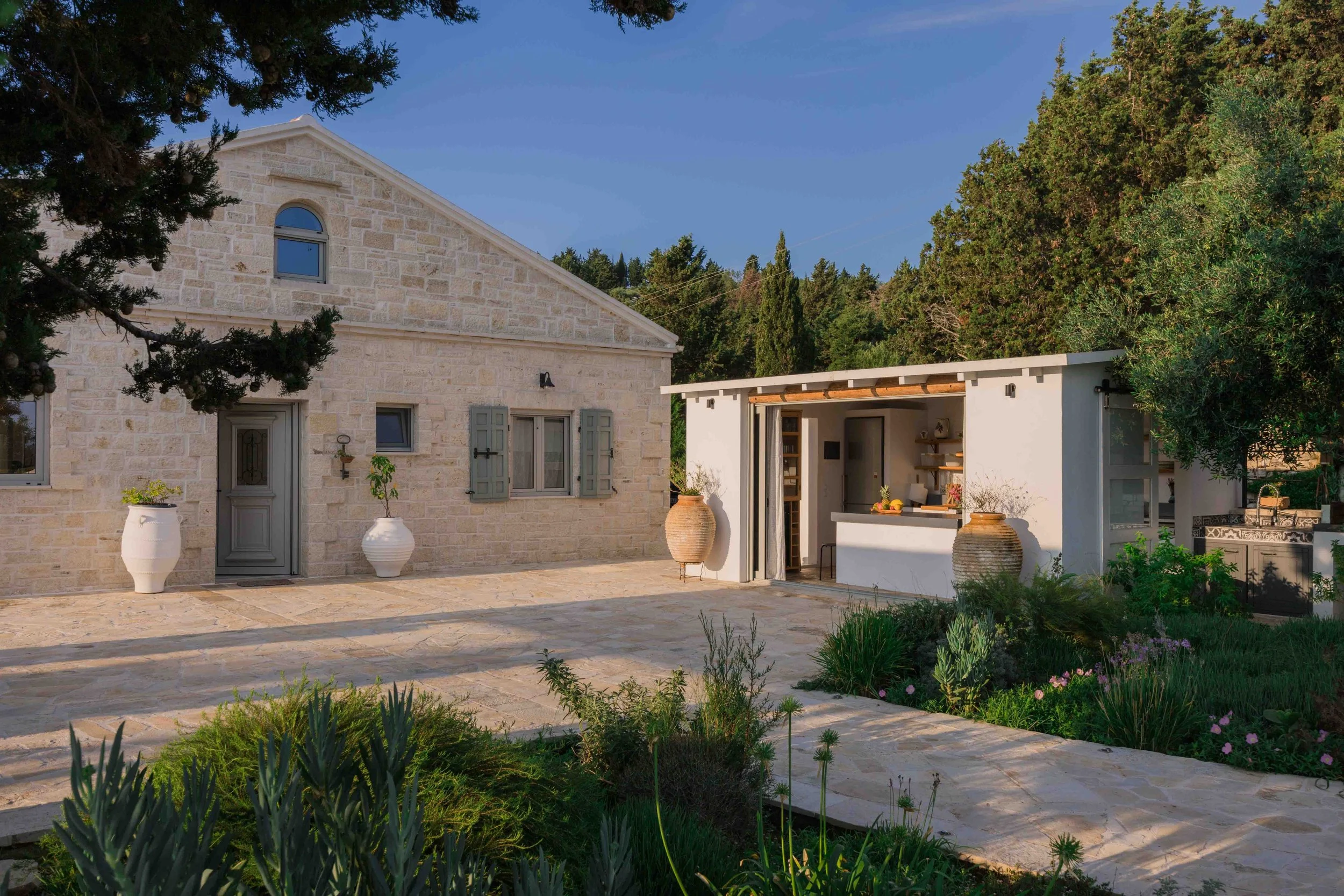 A stone house with window shutters and a door, a white outdoor kitchen area with shelves, two large pottery planters, and a garden with various plants and flowers, all under a clear sky surrounded by trees.