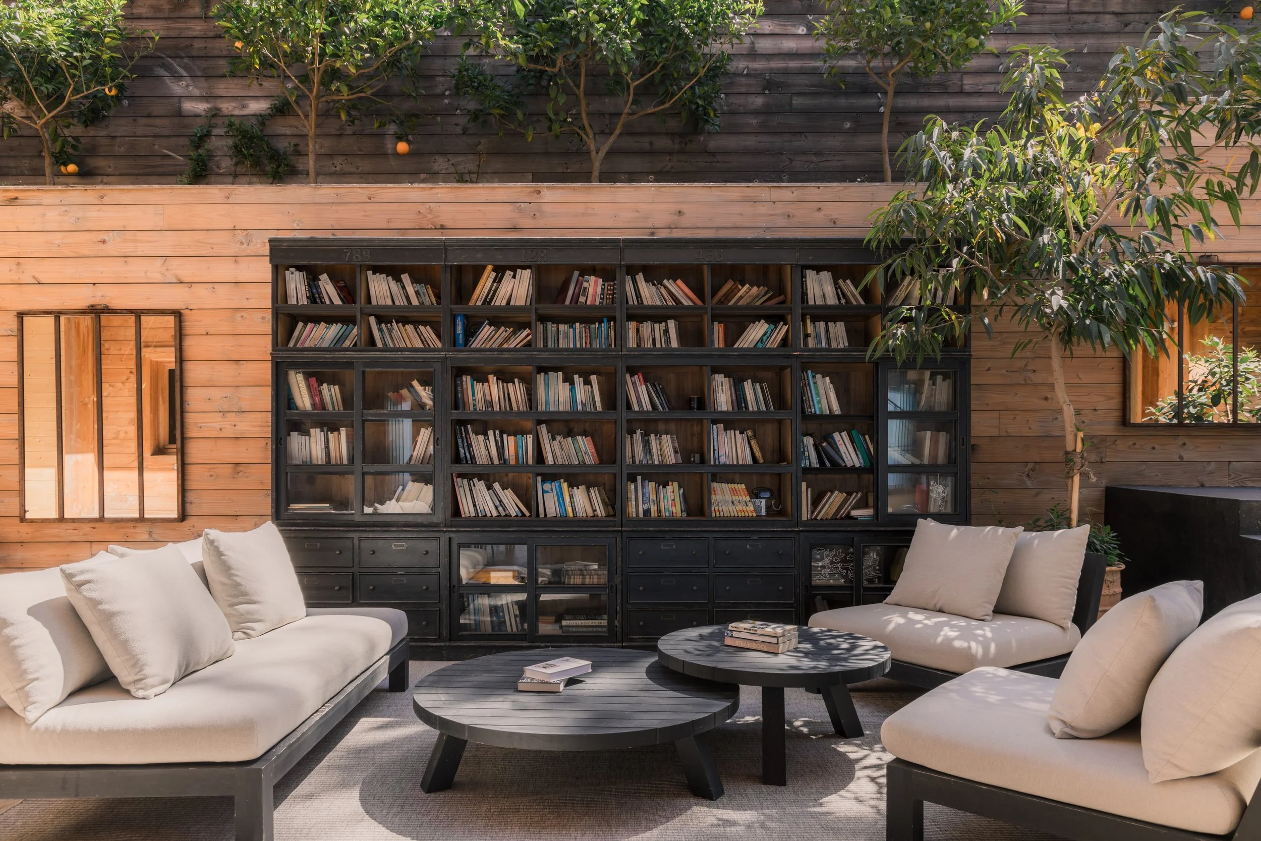 Outdoor living room with white sofas, black coffee tables, a large black bookshelf filled with books, wooden wall with greenery and orange trees, mirror on the wall, and potted plants.