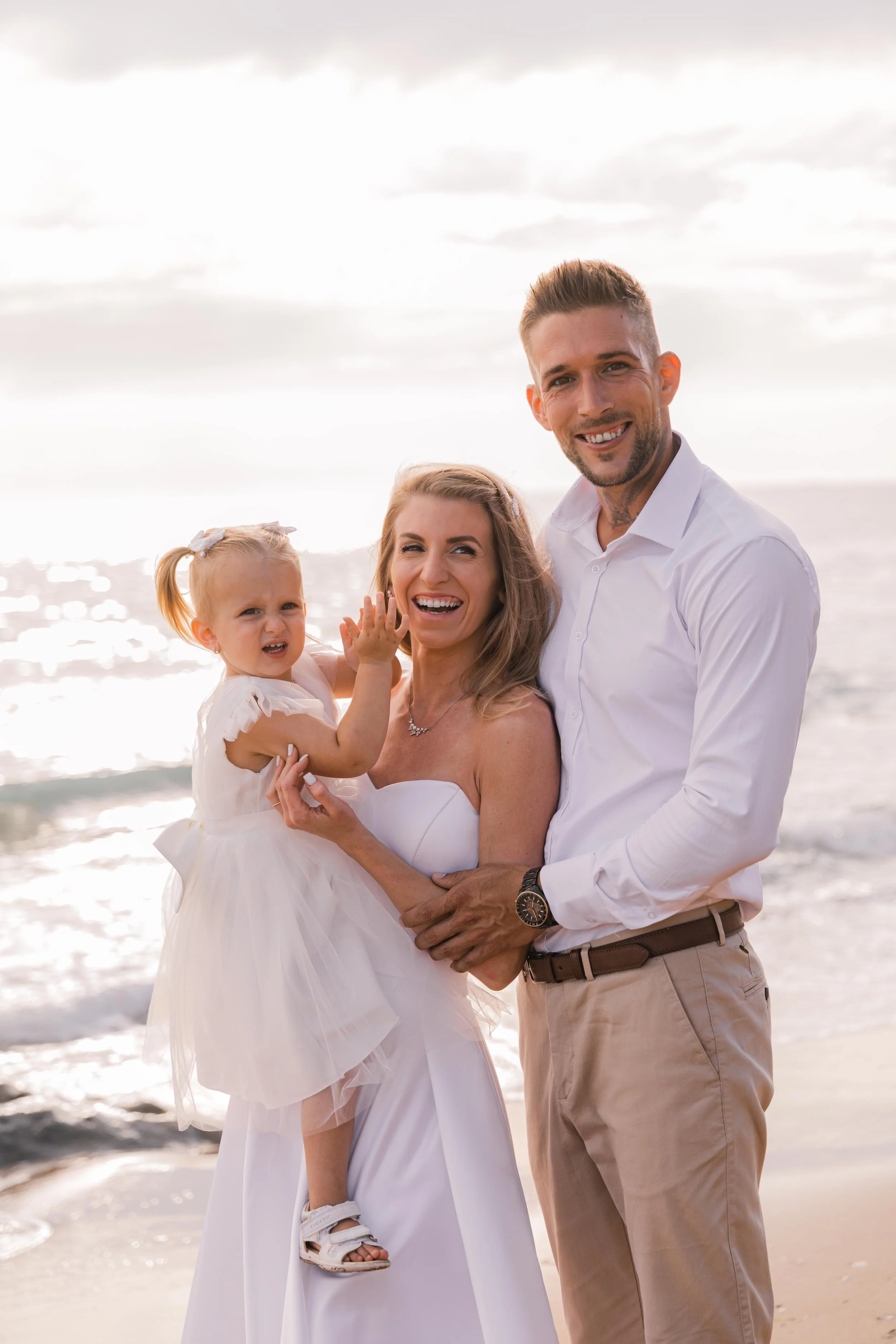 Family of three at the beach, smiling. Woman in white dress holding a young girl in a white dress, man in white shirt and beige pants standing next to them. Overcast sky, ocean in the background.
