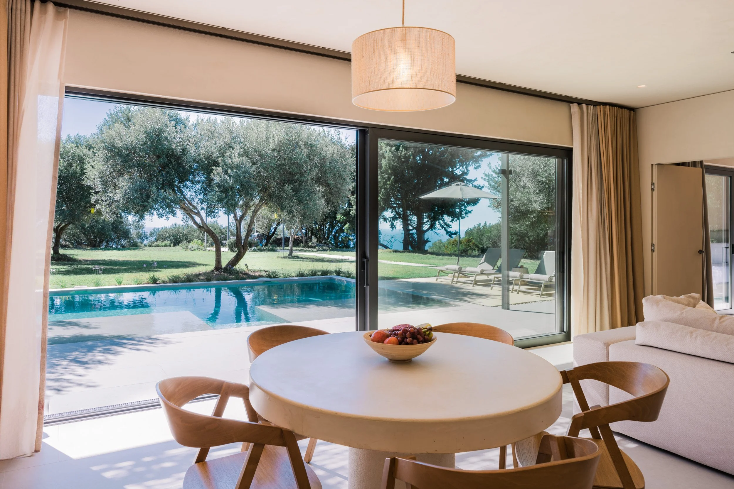 Modern living room with large glass sliding doors opening to a backyard with a swimming pool, lounge chairs, trees, and an umbrella