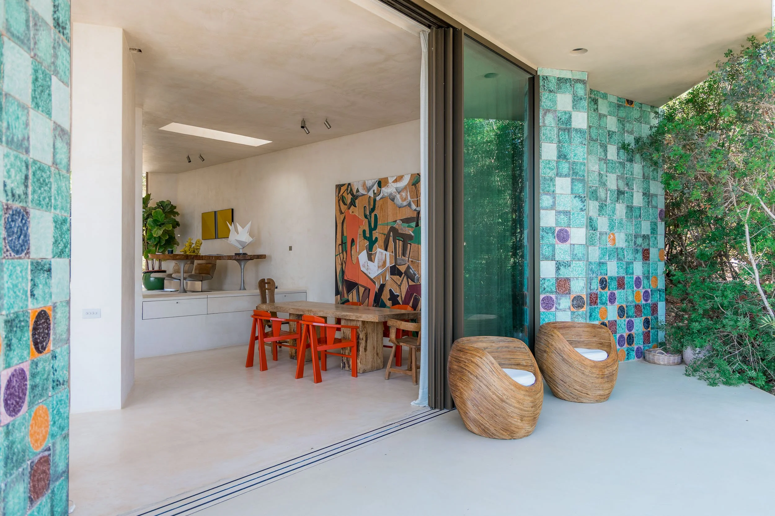 Interior view of a modern home with a sliding glass door leading to a balcony with two wooden chairs, colorful tiled wall, and a dining area with a rustic table and red chairs, decorated with abstract art and plants.