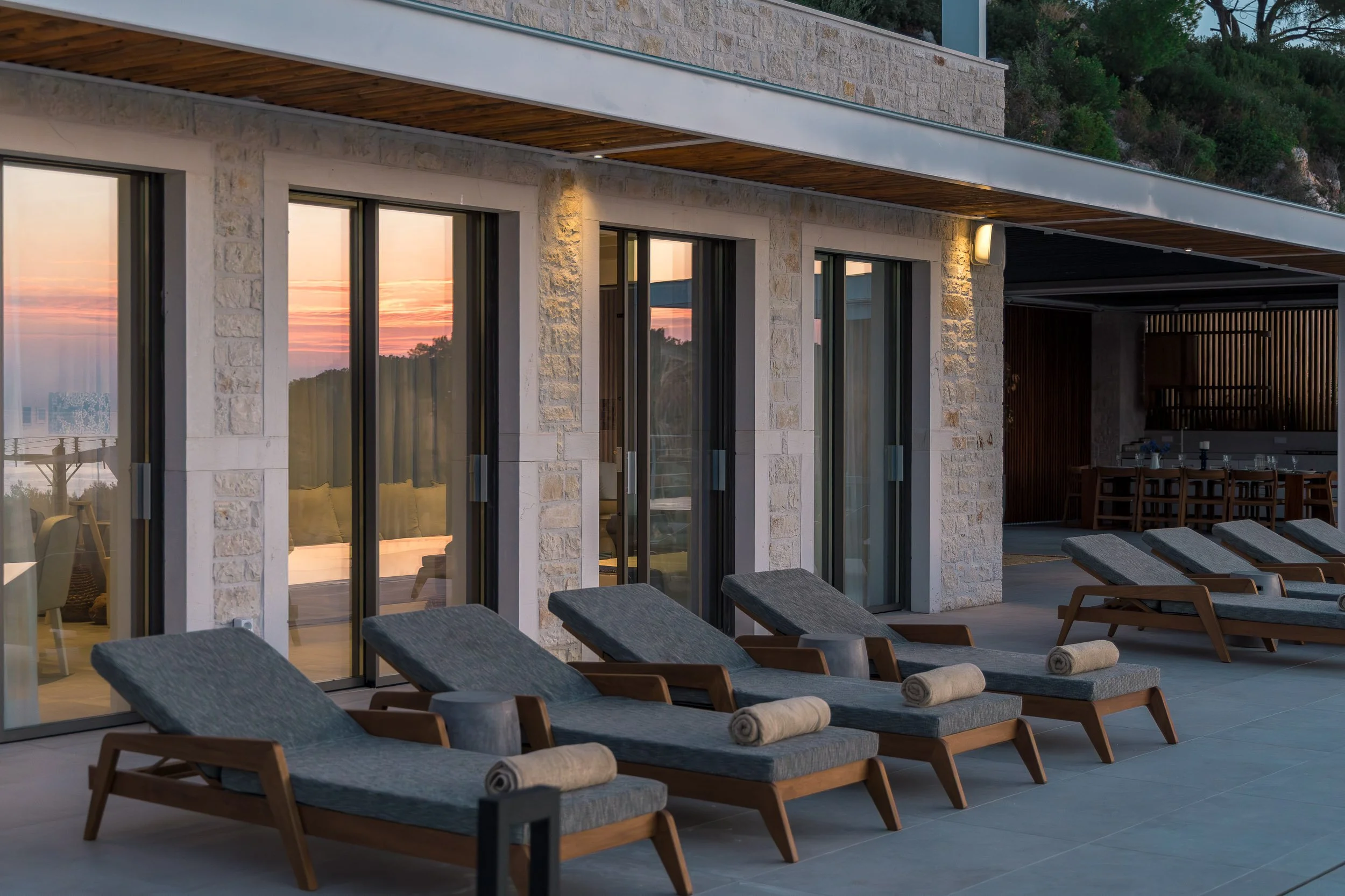 Outdoor terrace with lounge chairs and rolled towels, overlooking a sunset or sunrise with a view through large glass doors of an indoor lounge area.