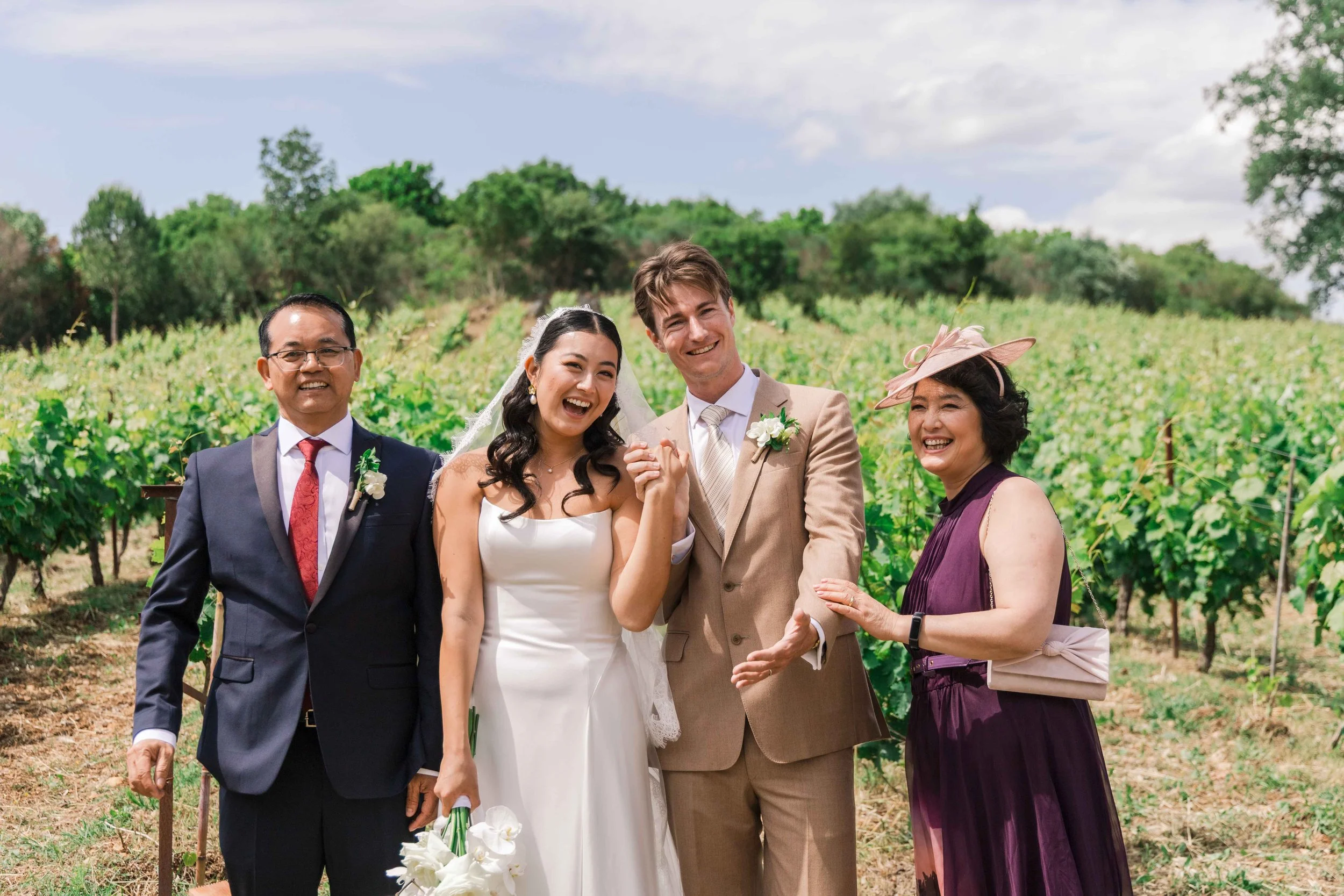 A wedding celebration with a bride and groom standing outdoors in a vineyard, smiling and holding hands, accompanied by three family members, against a backdrop of green vines and a partly cloudy sky.