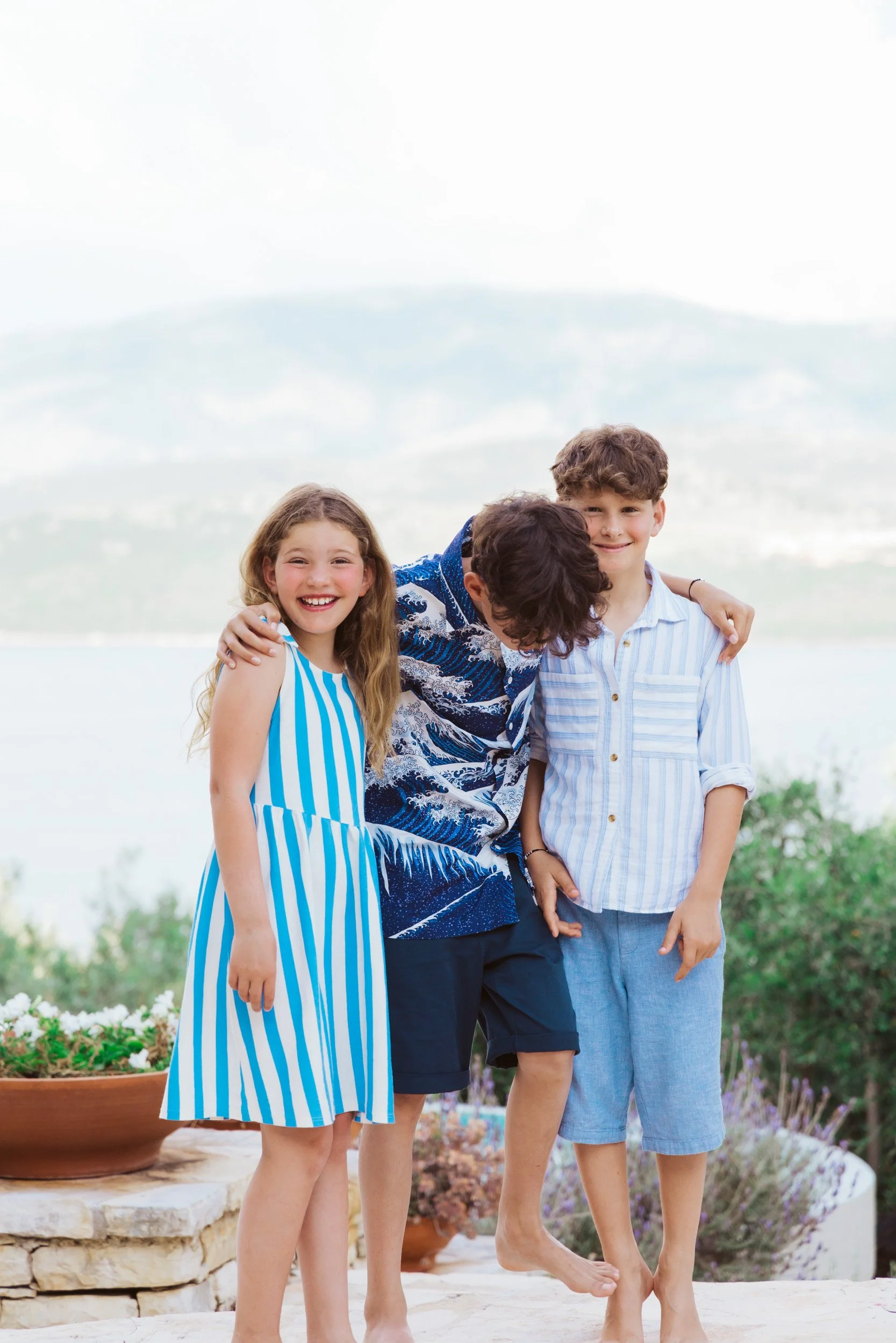 Three kids smiling and hugging outdoors with a lake and mountains in the background.